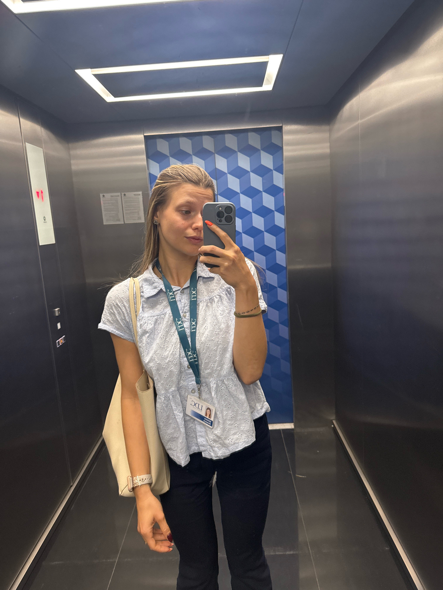 A blonde woman stands in an elevator and takes a selfie in a smartphone. She is wearing a light blue short-sleeved top, dark pants, and a beige tote bag over her left shoulder. A lanyard with an ID card is around her neck. The elevator walls are metallic, and the floor is black. The back wall of the elevator has a blue and white geometric pattern. The ceiling has a rectangular light fixture. The woman's fingernails are painted red. The scene is devoid of any context that would suggest a specific location within the city of Sofia, Bulgaria.