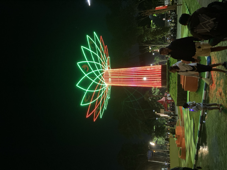 The image depicts a public park scene at night, centered around the Sidoarjo Monument (Monumen Jayandaru) in Sidoarjo, Indonesia. The dominant feature is a tall, stylized tree-like monument. Its cylindrical trunk is vertically striped with red LED lights, while the canopy is formed by layers of neon-green and red light strips arranged in a lotus flower pattern, with stylized shrimp or fish motifs illuminated in gold within the green petals.
In the immediate foreground, a child wearing light-colored clothing stands facing the monument, holding a glowing blue toy. To the child's right, a woman in a white long-sleeved shirt and dark skirt stands holding a white bag. Next to her, a man in a dark hoodie and light shorts stands with a neutral expression. Further right and slightly closer to the viewer, a person in a dark hoodie with "ALAN WALKER" text on the back is crouched or seated on the ground, facing away from the camera.
The ground comprises wet paved walkways and segments of green grass, bordered by a white and blue painted curb. In the mid-ground, green landscaped mounds and orange-colored concrete planters are visible. Various indistinct figures are scattered throughout the park, some walking, others congregated. A red tent structure is present to the left of the monument. Trees line the background, and a large group of motorcycles is parked in the distance. The sky is dark, indicating nighttime conditions.