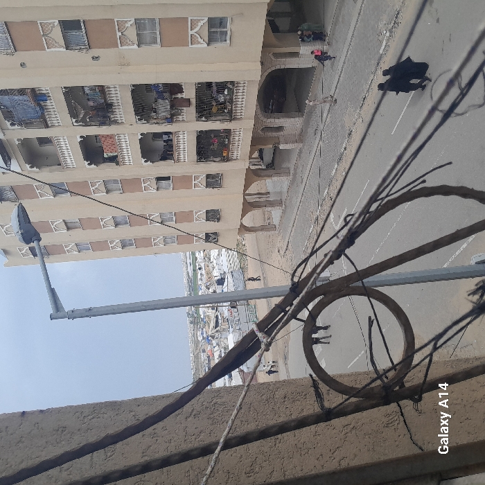 An elevated view in Khan Yunis, Palestinian Territory, captures a street, residential building, and a distant displacement camp under an overcast sky. In the foreground, a textured light-colored wall on the left is partially obscured by multiple dark electrical wires, including a prominent circular coil. "Galaxy A14" is visible in the lower-left corner.
The midground features a paved road with a lighter-colored sidewalk. A tall, grey metal street lamp with a modern fixture stands centrally. To the right, a six-story residential building with a light beige facade, brown vertical strips, and ornate white window frames and balcony railings dominates. Laundry hangs on several balconies. The ground floor is characterized by a series of arched open spaces.
On the street, two individuals dressed in dark abayas walk away from the viewer in the bottom right. Further along the street, a solitary figure in light clothing walks near the building's base. A small group, including a child in pink, walks closer to the building's arched ground floor, and another small group is visible on a dirt path to the left.
The background reveals an extensive area covered with numerous white tents, indicative of a large displacement camp. Beyond the camp, other urban structures and sparse vegetation are visible under a uniformly light grey sky.