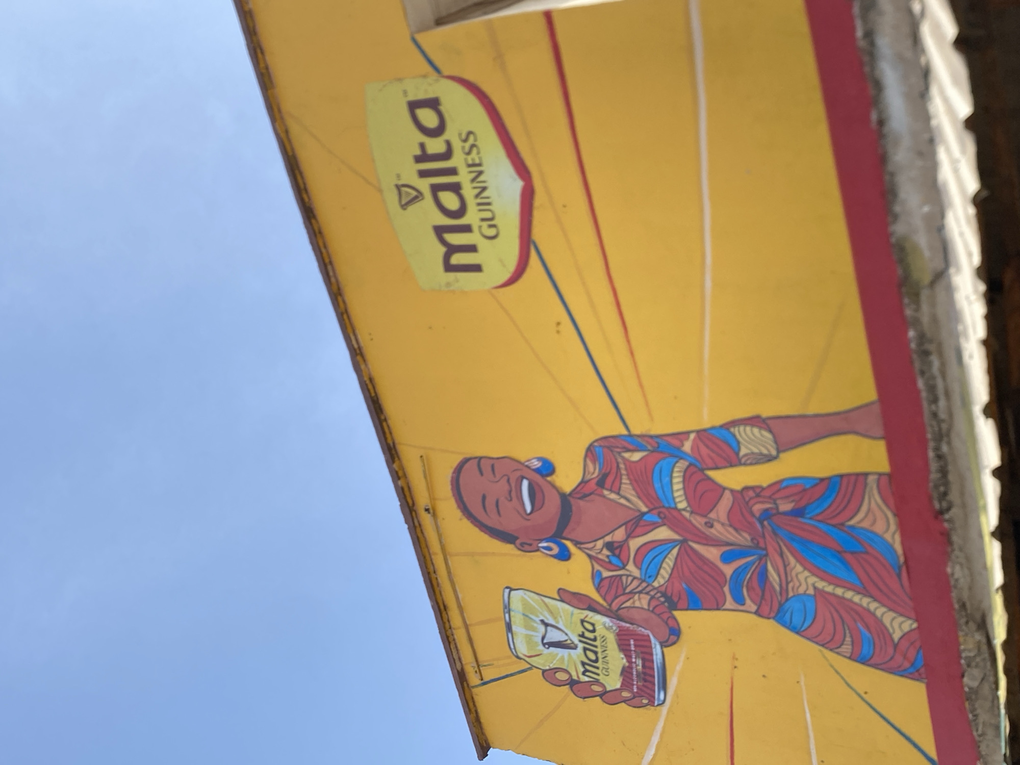 The image shows a partially visible Malta Guinness advertisement located in Bukuru, Nigeria. The advertisement is painted on a yellow background. The advertisement features a smiling woman wearing a patterned dress with red, blue, and yellow colors, along with blue earrings. She is holding a can of Malta Guinness. The sky is visible in the background. The sign appears to be attached to a weathered building or structure.