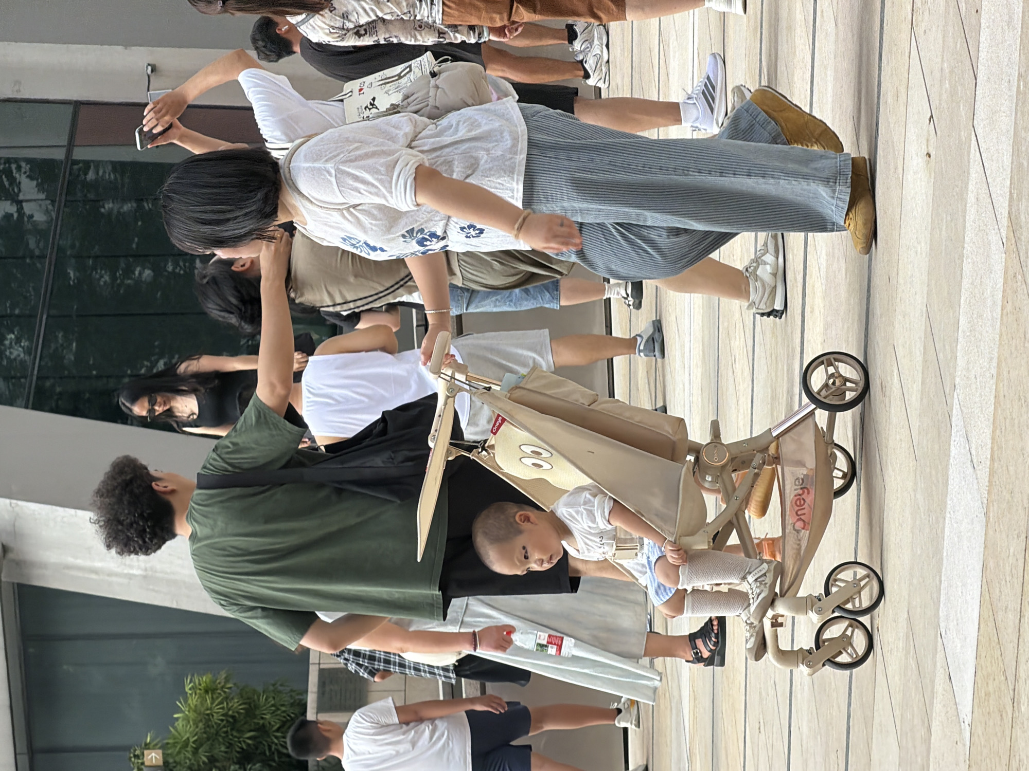 In Yuzhong District, China, an outdoor scene unfolds with several individuals present. In the foreground, a young child sits in a tan stroller, wearing a white t-shirt, blue shorts, and white socks with sneakers. The stroller is a prominent feature, with a stylized face design on its fabric. To the right of the stroller, a woman with short black hair, wearing a white patterned t-shirt and light blue striped pants, is captured mid-stride, holding the stroller's handle. She has a light-colored bag slung over her shoulder.
Behind the stroller and to the left, a man with curly dark hair, dressed in a green t-shirt and dark shorts, stands with his back mostly to the camera. He carries a black bag over his shoulder. Further to the left, a boy in a white t-shirt and blue shorts is also visible.
In the background, other individuals are present, including a woman in a black tank top and sunglasses, and another person holding a smartphone to take a picture. The environment appears to be an urban outdoor space with paved ground and a modern building with large glass windows in the background. The lighting suggests daytime.