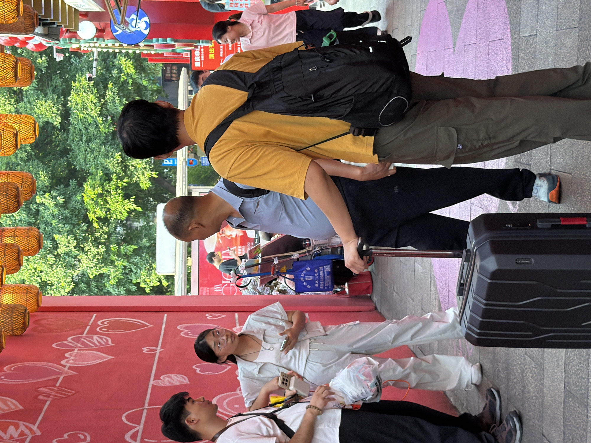 In Yuzhong District, China, a group of people are gathered on a pedestrian walkway. A young woman in white overalls and glasses stands near a young man in a pink t-shirt, both holding mobile phones. Behind them, an older man in a blue shirt faces away from the camera, while another man in a yellow t-shirt with a black backpack also faces away. In the foreground, a black suitcase rests on the ground. The environment features a red wall adorned with heart-shaped drawings and overhead decorative lanterns. Trees and buildings are visible in the background.