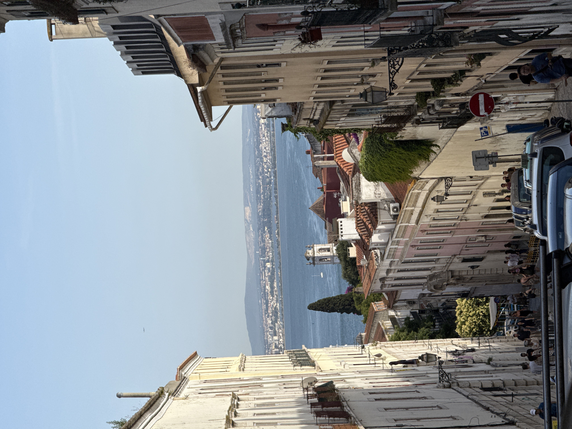 This image depicts a view down a steep, narrow street in Lisbon, Portugal, leading towards a body of water, likely the Tagus River, with a cityscape and hills in the distance. The street is lined with historic buildings, characterized by their colorful facades, balconies, and tiled roofs.
On the left, a tall, cream-colored building with arched windows and ornate balconies dominates the foreground. A satellite dish is visible on one of its balconies. Further down the street, on the right, buildings with beige, pink, and off-white facades are stacked closely together, featuring numerous windows and more balconies. Some buildings have ivy climbing their walls, adding a touch of greenery.
The street itself is paved and appears to be a pedestrian area, with a number of people walking down it. Some are dressed in casual summer clothing. Cars are parked along the sides of the street, including what appears to be a taxi. Traffic signs, including a "no entry" sign and a parking sign, are visible.
In the middle ground, the view opens up to the expansive blue water. A tall, slender evergreen tree stands prominently near the water's edge. A bell tower with a white facade and a clock face rises above the buildings, suggesting a church or cathedral. A white sailboat is visible on the water.
The background shows a sprawling cityscape on the opposite side of the river, with buildings and structures stretching towards the horizon, framed by distant, hazy blue hills. The sky is a clear, bright blue. An airplane is visible as a small dot high in the sky. The overall atmosphere is one of a vibrant, sunny day in a historic European city.