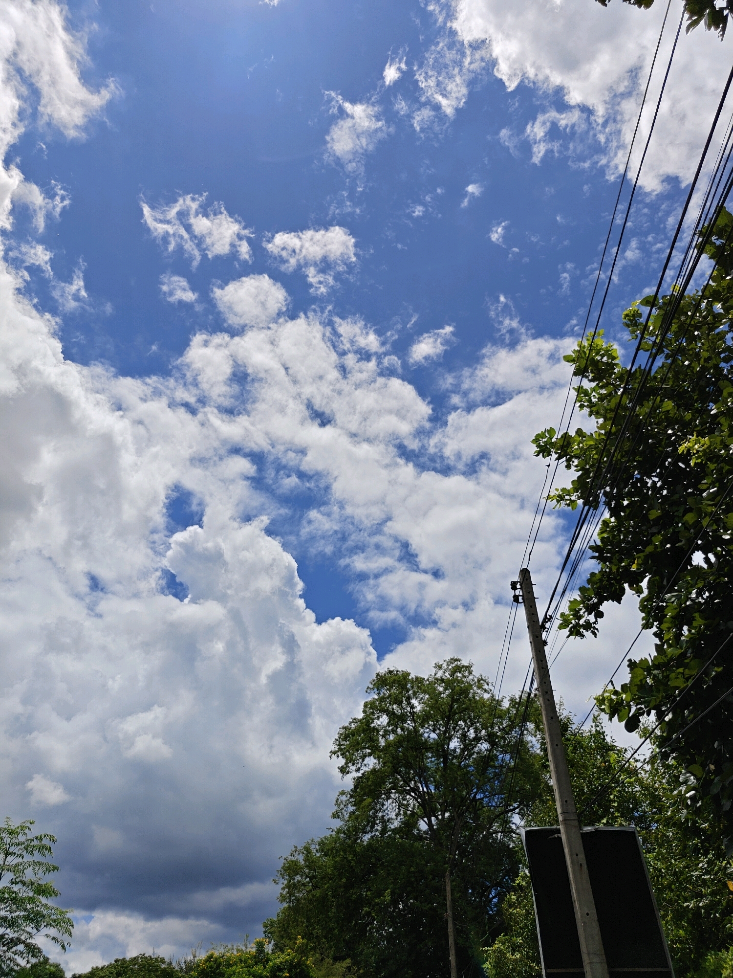 The image depicts a view looking upwards at a bright blue sky filled with white and grey cumulus clouds. In the foreground, to the right, a wooden utility pole supports several black electrical wires that extend diagonally upwards. Adjacent to the pole, lush green trees with dense foliage are visible, partially obscuring the sky. The lower portion of the image shows the tops of more trees and a dark, rectangular object, possibly a sign, attached to the utility pole. The overall scene suggests an outdoor environment in a tropical location. The image was captured in Mu Si, Thailand, as indicated by the surrounding vegetation and typical utility infrastructure found in the region. There are no people or animals present in the image.