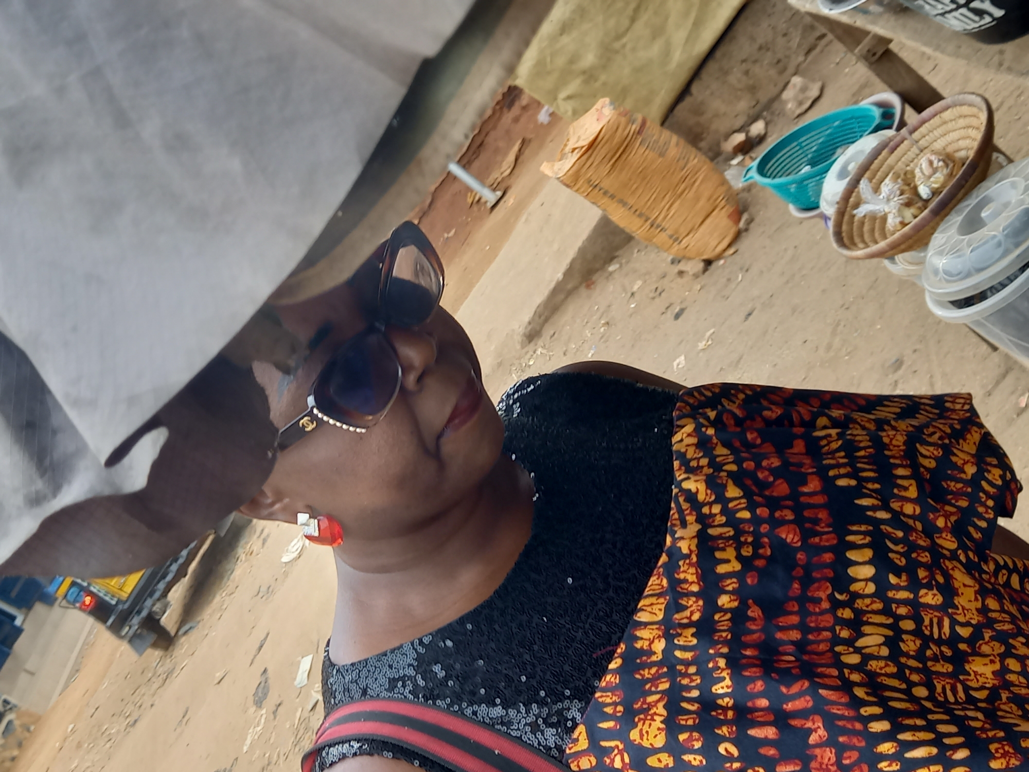 The image presents a close-up, angled portrait of a woman in an outdoor market setting, likely in Onitsha, Nigeria. The woman, of medium complexion, is seen looking slightly upwards and to her right, wearing large, stylish sunglasses with a gold-colored detail on the arm, vibrant red lipstick, and prominent red earrings. She is dressed in a black top or dress heavily adorned with sequins. A richly patterned fabric, featuring orange and yellow abstract designs on a dark background, is draped over her left arm or shoulder, adding a pop of color and cultural flair.
The background depicts a dusty, uneven ground typical of an outdoor market. Various items are scattered on the ground, including a large, light-brown sack, a bright turquoise plastic basin, and a woven straw basket containing small, light-colored objects, possibly shells or crafts. Several clear plastic containers with lids are also visible. In the upper left background, a white tarp or sheet provides shade, beneath which parts of a yellow vehicle, possibly a truck or bus, can be seen. The scene is brightly lit, indicating daytime. The woman appears calm, possibly smiling slightly, suggesting she is either posing or paused in her activity. No explicit text is clearly visible in the image.