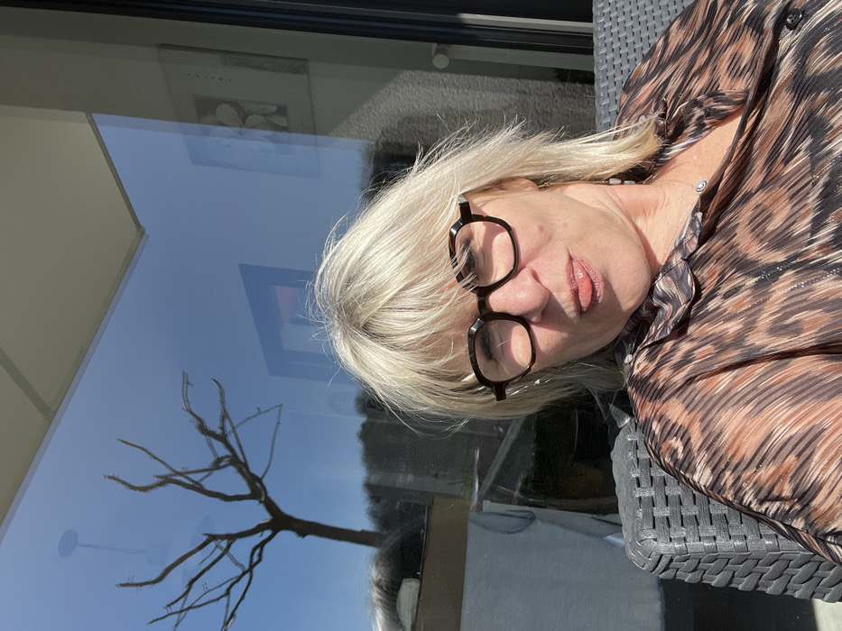 A blonde-haired Caucasian female, wearing dark hexagonal glasses and a brown-and-black patterned blouse, is depicted seated outdoors in a dark grey woven chair under direct sunlight in Chambœuf, France. She is making a playful facial expression with her tongue slightly extended. Behind her, a large glass window reflects a bare-branched tree against a clear blue sky, distant foliage, and interior elements including a framed picture and a ceiling light fixture.