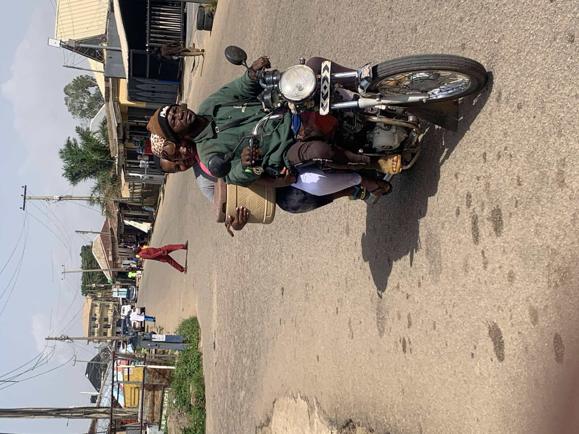 In the city of Jos, Nigeria, on a paved road under a bright sky, three individuals ride a motorcycle. The driver, a man wearing a green jacket and a beanie, navigates the vehicle. Behind him, two passengers, likely women, are seated. One passenger wears a pink shirt and a patterned headscarf, while the other is in a grey shirt. The woman in the pink shirt holds a beige cooler box. The motorcycle itself is dark red and displays decorative elements on its front. The road shows signs of wear and tear, with patches and discoloration. Buildings line the street, some appearing to be shops with visible wares. Electrical wires crisscross overhead, a common sight in urban Nigerian landscapes. In the background, a person in a red outfit walks across the road, and other vehicles are present further down the street. The scene suggests a bustling everyday moment in Jos.