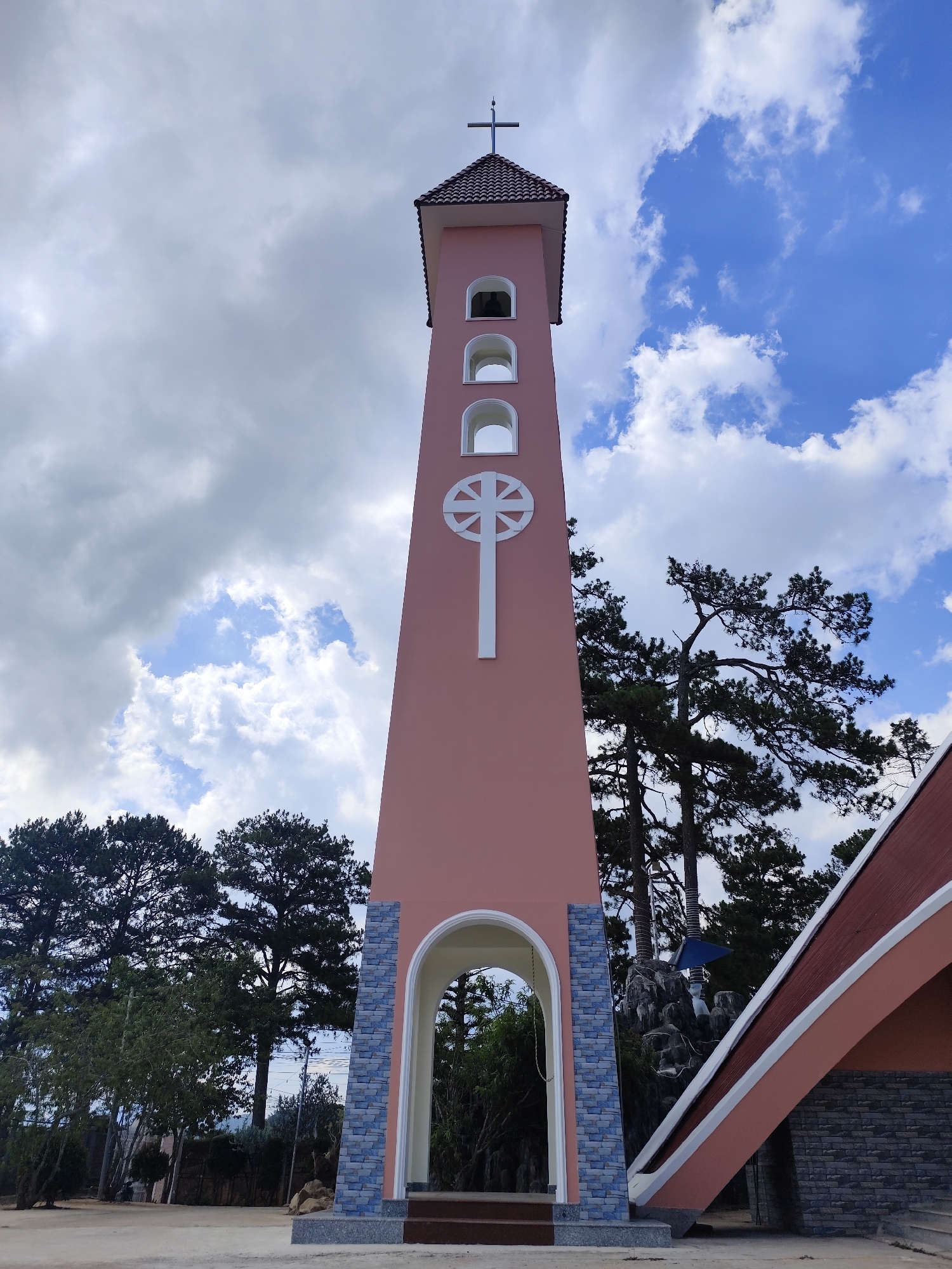 A ground-level, medium shot of a pink, four-story bell tower of a Catholic church, located in the city of Đa Thọ, Vietnam. The tower has a tiled, pointed roof with a cross on top. The second and third stories from the top are arched, containing bells. A white stylized cross is emblazoned in the middle of the tower. The base of the tower is an arched entryway framed by blue-gray brick. The ground is light colored cement. Trees are visible in the background, as well as blue and white clouds in the sky. The corner of a low building with a sloped roof is visible in the lower right corner.