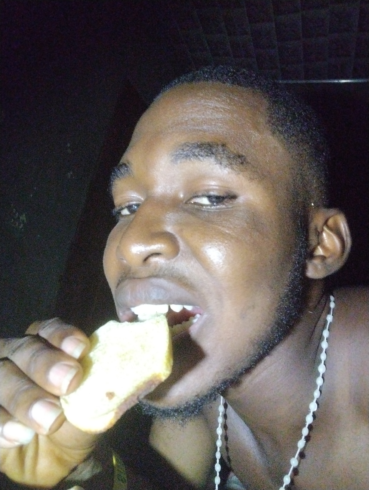 A close-up, dimly lit image captures a person, likely male, with dark skin, short dark hair, and a short beard or stubble. The individual is actively eating, holding a piece of light-colored food, resembling bread or cake, up to their mouth and taking a bite. A white beaded necklace is visible around their neck, draped over their chest. The background is dark and appears to be an indoor setting, possibly a wall or paneling, with a faint grid-like texture visible in the upper right. The low light conditions suggest either nighttime or an enclosed environment. The person's eyes are open and directed slightly towards the camera, with a focused expression as they eat. No text is visible in the image. The context provided for the location is Ihiagwa, Nigeria.