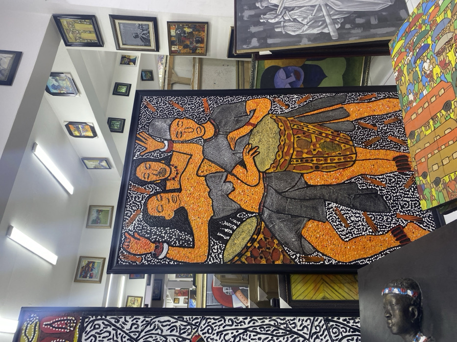 This image depicts an interior view of an art gallery or studio located in Alaguntan, Nigeria. The central focus is a large, vertical framed painting rendered in a textured, pointillistic style, featuring three stylized figures against a black background. The figures, depicted with orange-brown skin tones and black outlines, include two female figures with raised arms, one wearing a necklace, and a male figure in a grey tunic and cap actively playing a patterned drum. A second drum is partially visible to the left of the male figure.
The gallery space features white walls and ceiling illuminated by multiple fluorescent strip lights. Numerous framed artworks of varying sizes and styles, including landscapes and abstract pieces, are displayed on the walls. In the foreground, to the left, a portion of another large vertical painting with black and white abstract patterns is visible. A dark-colored bust or sculpture of a head, marked with red, white, and blue stripes on its forehead, rests on a dark surface in the immediate foreground. To the right, several paintings are stacked or leaning, including a grayscale work depicting multiple figures, a stylized human face in blue and green hues, and a colorful abstract composition. The floor is light-colored.