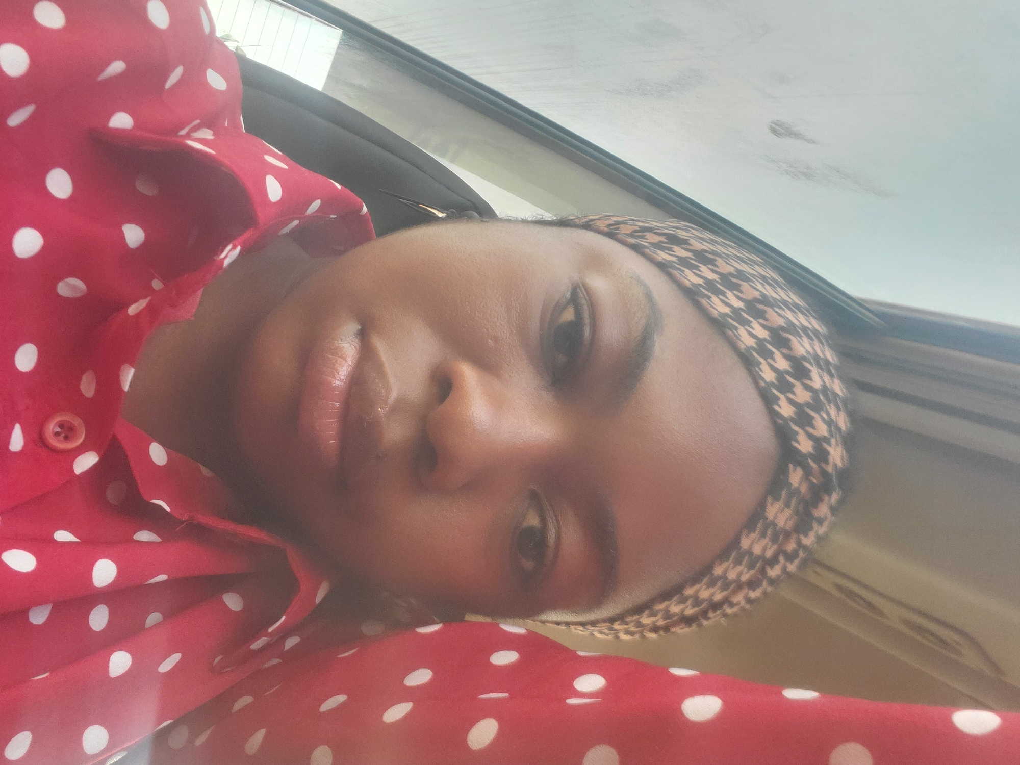 A close-up, medium shot shows a young Black woman from the chest up, facing forward and slightly to the left. She is wearing a red, polka-dotted button-down shirt and a patterned headscarf. The background is blurred and appears to be the interior of a vehicle, with a window on the left side showing a faint reflection of what might be a sky. The lighting is soft and diffused. The woman's facial expression is neutral.