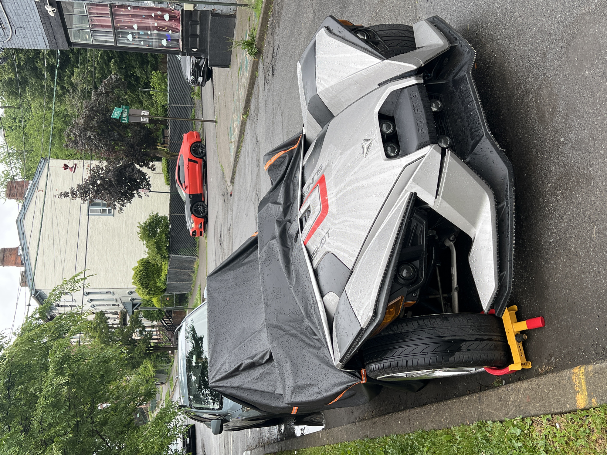 The image depicts a street scene in the City of Newburgh, United States of America. A Polaris Slingshot three-wheeled vehicle is parked on the side of the road. The vehicle is partially covered with a dark-colored tarp. The front driver's side wheel of the vehicle is immobilized by a yellow wheel clamp with red handles. In the background, there is a street sign indicating "First Lander St," along with other parked vehicles including a red sports car and a black vehicle. Buildings and trees line the street. The road surface appears wet, possibly due to rain.