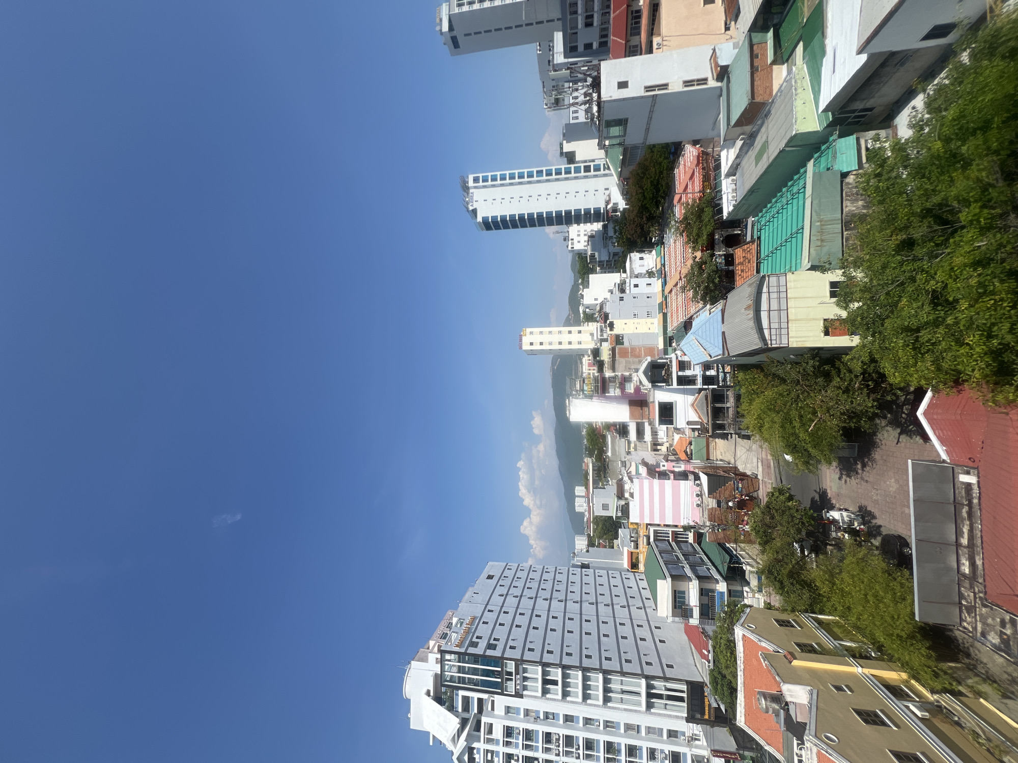 A high-angle, vertical, medium shot captures a dense urban landscape under a clear, bright blue sky in Nha Trang, Vietnam. The composition is dominated by a variety of buildings, ranging from tall, modern high-rises to smaller, older structures with colorful roofs.
In the foreground and to the left, a large, white, multi-story hotel with a prominent glass facade and blue accents stands out. Its signage reads "Moonlight Hotel." Adjacent to it are other buildings with varying architectural styles and colors, including a structure with pink and white stripes.
The mid-ground reveals a street lined with more buildings, many featuring green, red, and yellow roofs. Trees with lush green foliage are interspersed throughout the urban fabric, providing a contrast to the concrete and glass. A few vehicles, including a black car and what appear to be motorcycles, are visible on the street.
In the background, several more contemporary buildings rise, including a tall, slender white tower with blue windows. Further in the distance, mountains are visible under the expansive sky, with a few wispy clouds. The overall impression is a vibrant, bustling city scene on a sunny day.