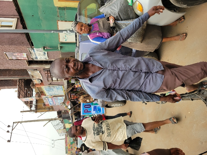 The image displays a bustling street scene in Onitsha, Nigeria, characterized by pedestrian traffic and commercial activity under an overcast sky. In the foreground, an adult male with a short, dark beard and shaven head walks towards the viewer, his gaze directed forward. He wears a dark blue-grey long-sleeved shirt, unbuttoned at the collar, and brown trousers, holding a metal object, possibly a mechanical component, in his right hand. To his immediate left, another adult male walks away from the viewer, wearing a cream-colored t-shirt with "BURBERRY" text, light grey shorts, and dark sandals. He is looking down at a smartphone in his left hand while carrying a black plastic bag in his right. To the right of the central figure, an adult female with a patterned headscarf, purple t-shirt, and dark brown apron stands beside a silver sedan, attending to items near the vehicle, including a large grey bag.
The background features multiple multi-story buildings of varying colors, including shades of brown, green, and yellow, lining the street. Numerous commercial signs are affixed to these structures, one prominent yellow sign on the far left reads "OGENE MAJMAJMA'S SUPER MARKET." Utility poles with overhead power lines are visible on the left, and a grey utility pole stands on the right. The ground is an unpaved or dirt-colored road surface. Several other individuals are partially visible in the midground and background, indicating a populated area. Vehicles, including a dark-colored car, are parked or moving within the scene.