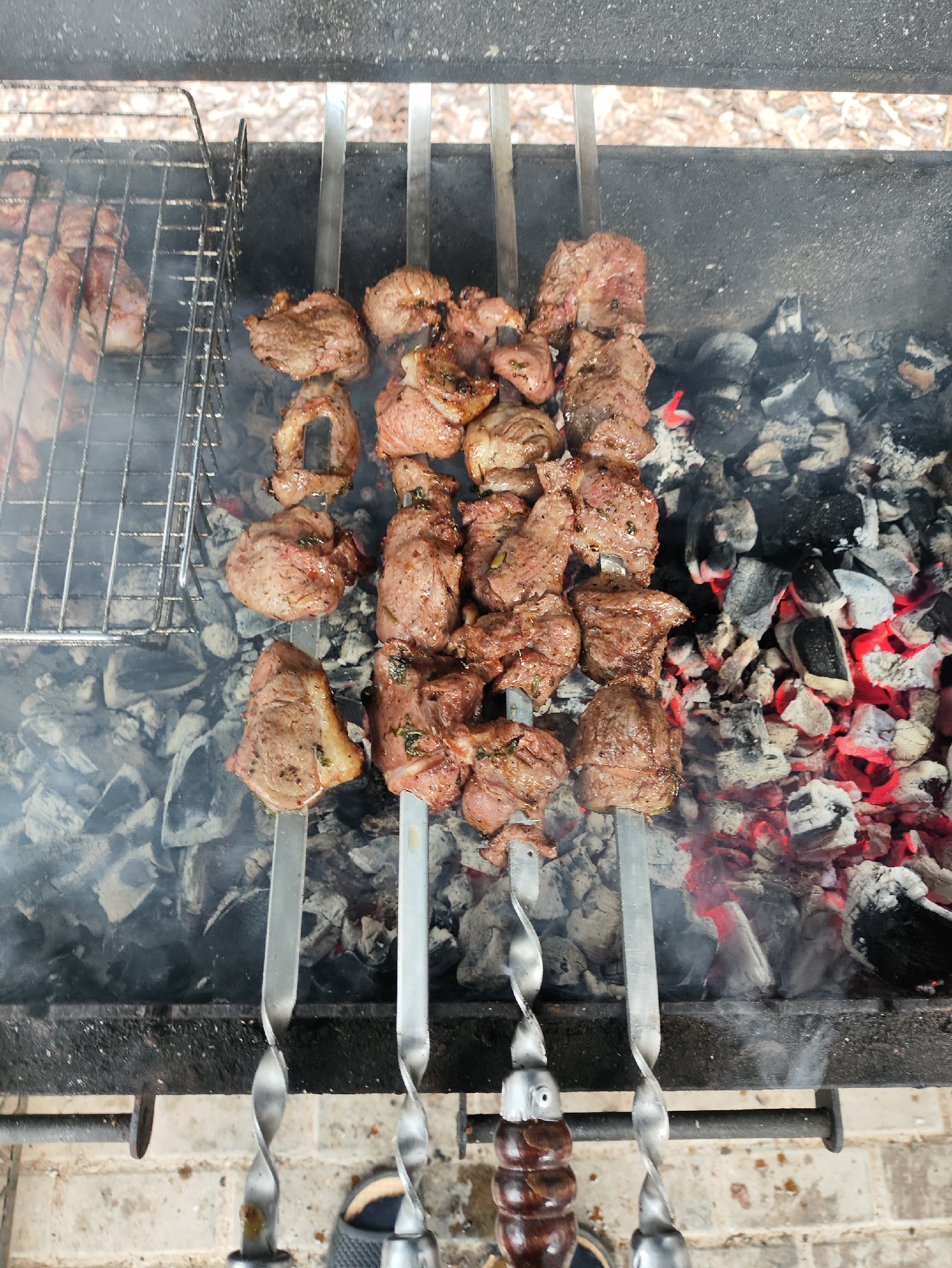 A top-down, medium shot captures skewers of seasoned meat being grilled over glowing red coals in a brazier. The skewers are arranged in a row, with the meat pieces varying in size and shape, some appearing to be lamb or beef. To the left, a wire grill rack holds more raw meat. Smoke billows up from the coals, partially obscuring the lower portions of the skewers. The brazier itself is set on a brick surface, with a textured dark metal surround. The background is blurred, showing a hint of foliage and what appears to be a light-colored gravel or dirt path. The overall scene suggests an outdoor barbecue setting, likely in a village or rural area given the context of Старое Мартьяново, Russia.