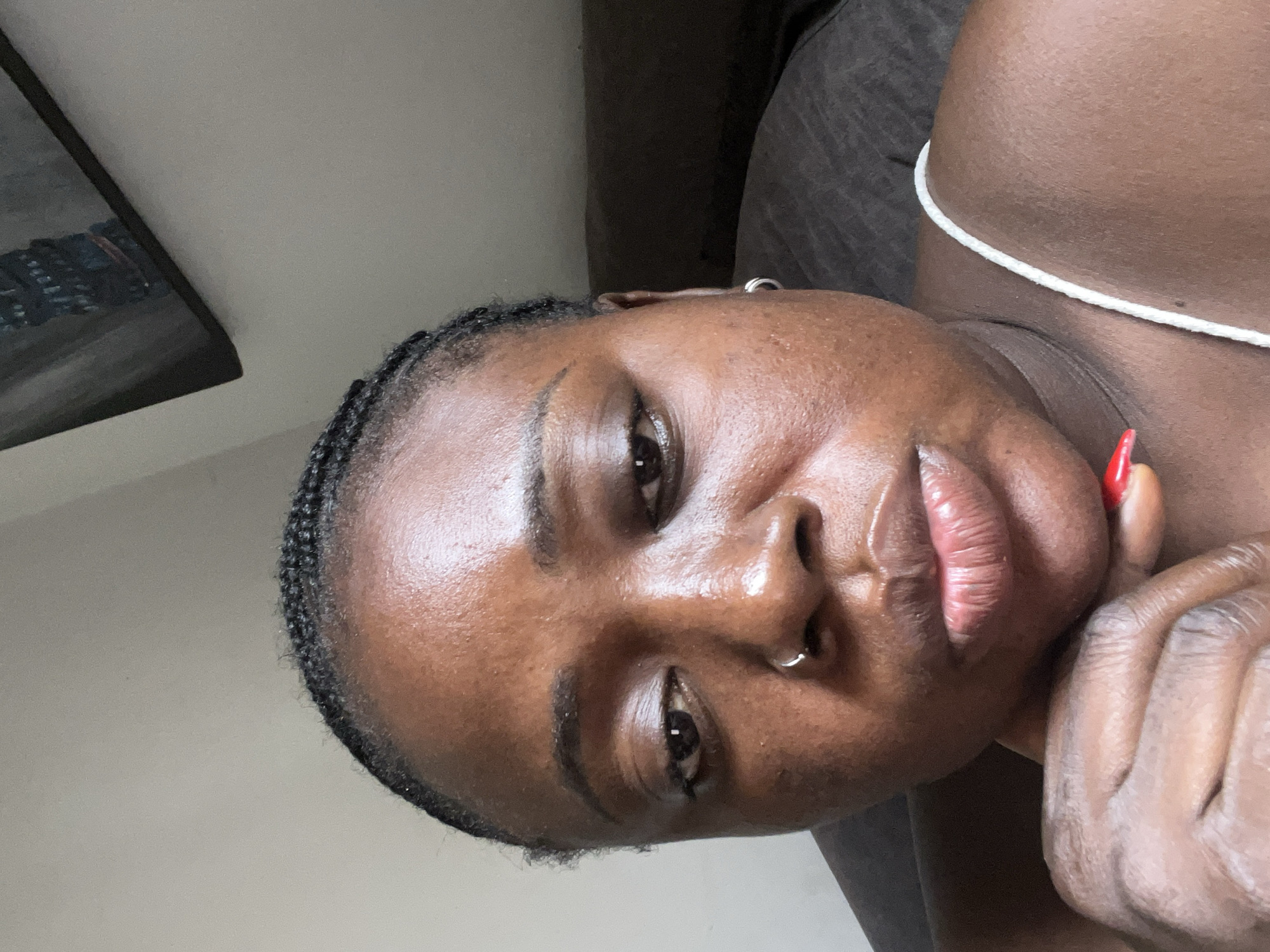 The image is a close-up selfie of a young Black woman. She is looking directly at the camera with a neutral expression. Her hair is styled in braids, and she is wearing a white strap top. She has a nose ring and a small hoop earring in her left ear. Her right hand is resting on her chin, and her fingernail is painted red. The background is blurred but appears to be an indoor setting with a wall and a piece of artwork on it. The lighting is soft, suggesting it might be natural light from a window. There is no indication of the specific location within Abuja, Nigeria, or any details related to the city's environment.