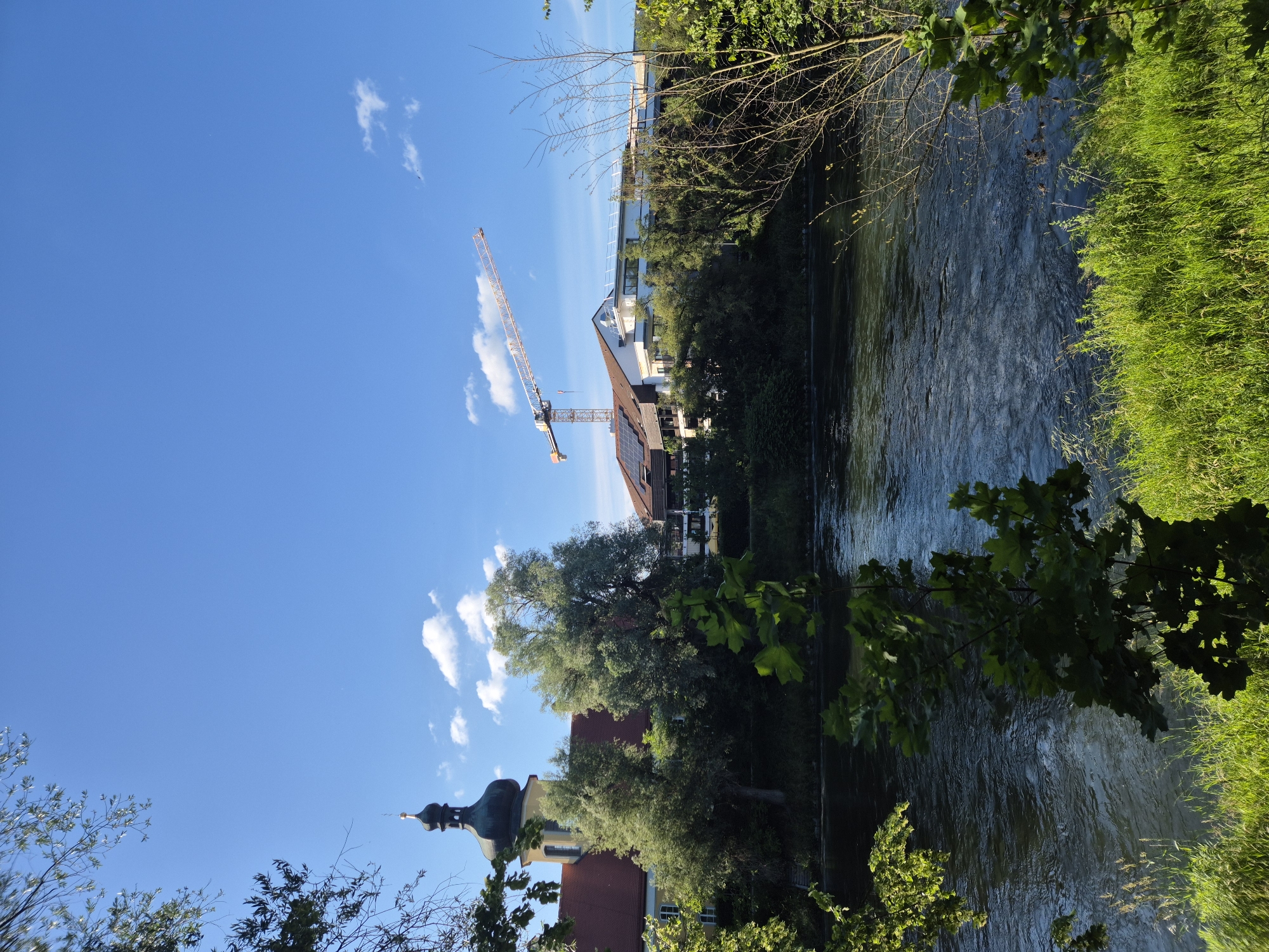 The image depicts a scenic view of the Isar river in Landshut, Germany. On the left bank, a building with a distinctive onion-domed tower, likely a church or monastery, is partially visible behind lush green trees. Across the river, on the right bank, a modern building with a white facade stands prominently. A tall construction crane is positioned over this building, suggesting ongoing development. The river itself is calm with gentle ripples reflecting the bright blue sky dotted with white clouds. The foreground is filled with vibrant green grass and foliage, framing the entire scene. The sun appears to be shining, casting shadows and illuminating the landscape.