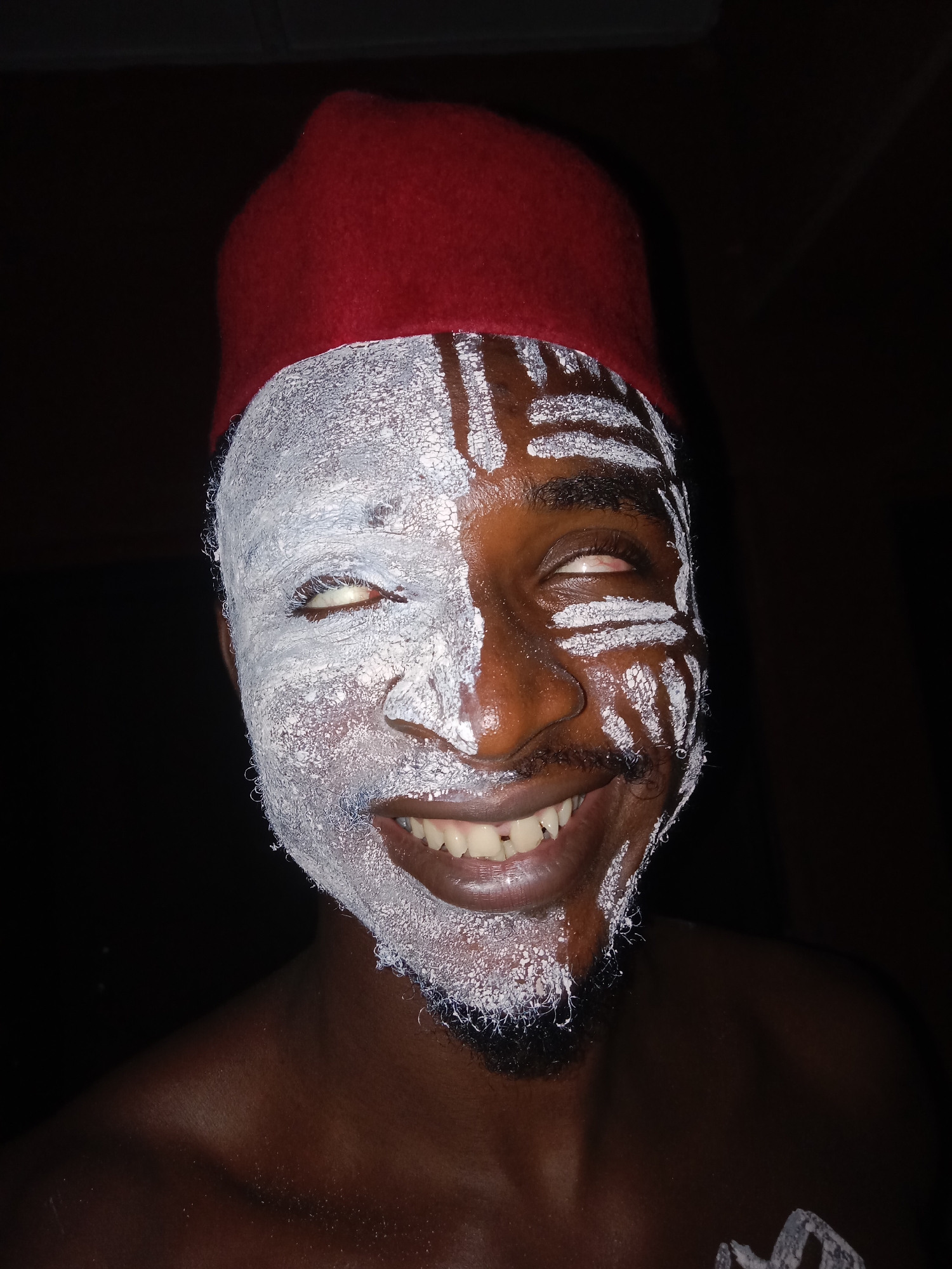 A close-up shot depicts a person wearing a red fez-style hat. Half of their face is covered in thick white paint, while the other half features white lines and patterns painted on the exposed dark skin. The person is smiling, revealing their teeth. Their eyes appear to be rolled upwards, showing mostly the whites. A partial beard is visible. The background is dark. The person appears to be wearing a cultural or ceremonial costume. Based on the fez hat and face paint, it may depict a performance or ritual related to the Igbo people. There is no information in the image confirming the location in the city of Onitsha, Nigeria.
