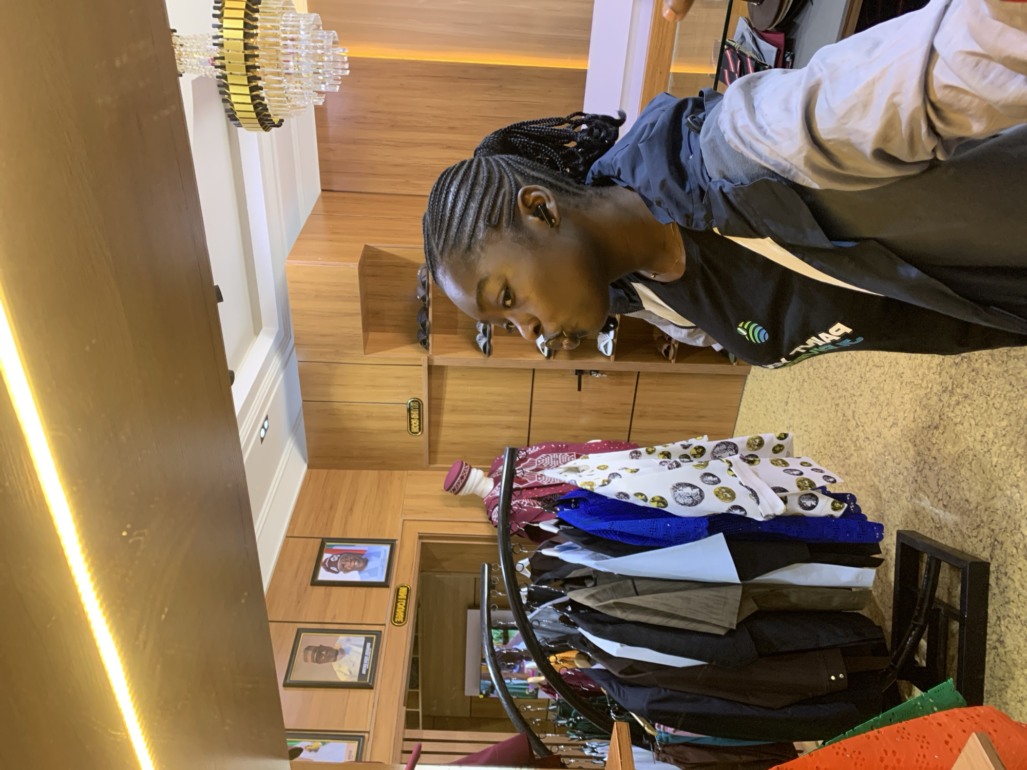 The image captures a young Black woman with braided hair styled in a ponytail, wearing a black t-shirt with a logo and a blue and gray hooded jacket. She is standing in what appears to be a clothing store. In the background, there are racks of clothing, including suits and traditional Nigerian attire on mannequins. Framed pictures of men in formal wear are displayed on the wall, along with signage. A chandelier hangs from the ceiling. The environment suggests an upscale retail space, possibly a tailor's shop or a boutique selling formal and traditional clothing. The woman's posture and expression indicate she is looking away from the camera, perhaps engaged in a conversation or observing something in the store. The lighting in the store is warm, emanating from overhead fixtures and accent lights. The floor has a speckled texture. The overall scene depicts a commercial setting within a city, possibly Gura Top, Nigeria, given the context provided.