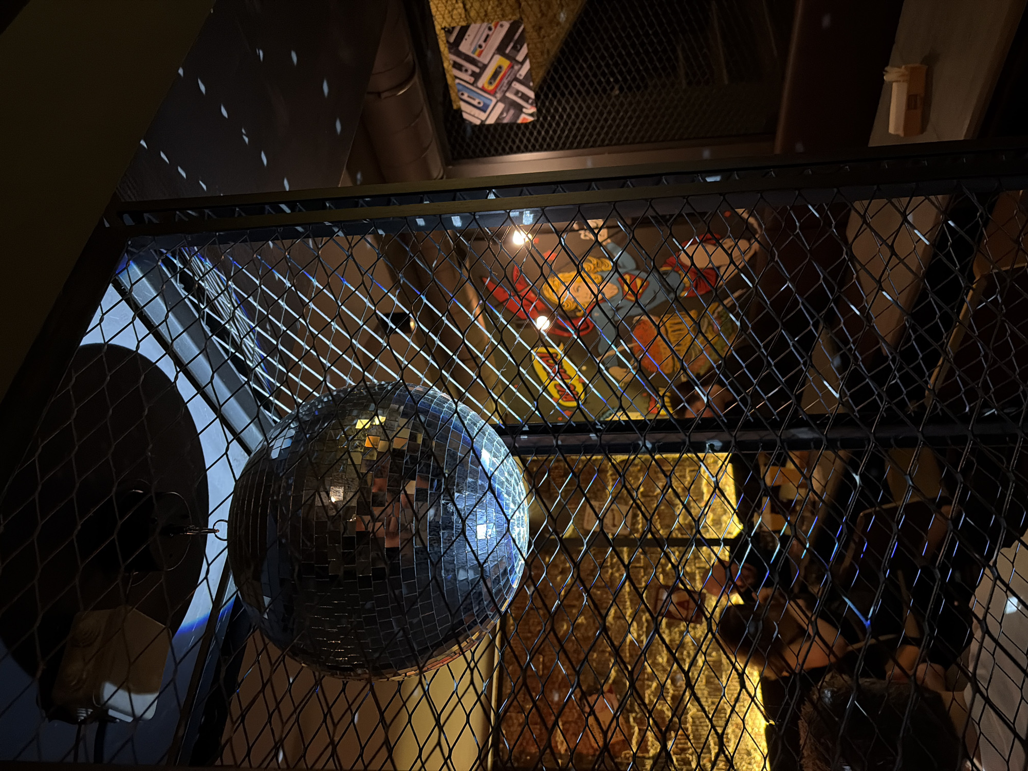 An indoor, dimly lit scene features a disco ball suspended by a chain, reflecting light in a room with a brick wall and a metal mesh partition. Through the mesh, several people are visible in what appears to be a seating area, possibly a bar or restaurant. A decorative item depicting cassette tapes hangs on the wall in the background, and a small, yellow dispenser is present on a surface to the right. The overall atmosphere suggests a casual, social setting. The location is specified as Minsk, Belarus.