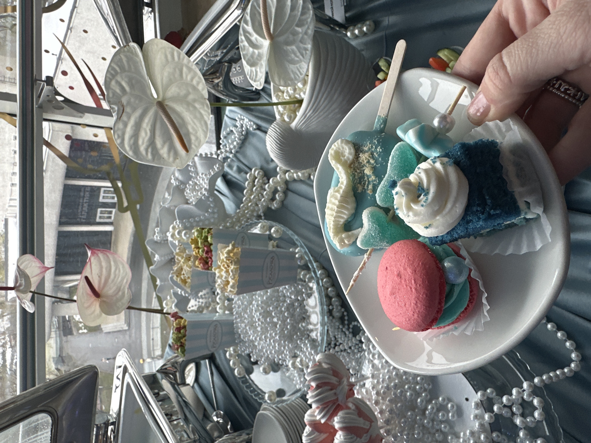 The image, taken from an overhead and tilted perspective, captures an elegant dessert display, likely part of a celebration or party set up indoors during the daytime. A hand, with neatly manicured nails and a ring on one finger, holds a small white plate filled with an assortment of blue and pink desserts. On the plate, there's a blue cake pop or treat on a stick, decorated with a white seahorse and sprinkles, alongside blue gummy candies. A piece of blue cake with white swirled frosting and blue sprinkles, and a bright pink macaron with light blue filling in a paper cup, complete the plate's offerings.
The dessert table is lavishly adorned with a light blue and white color scheme. White pearls and beads are scattered abundantly over a light blue fabric surface, possibly a tablecloth. Elegant white anthurium flowers, some with delicate pink edges, are arranged in vases, enhancing the sophisticated aesthetic. A white, textured, shell-like or wave-shaped decorative object is visible among the pearls.
In the background, small, vertically striped blue and white popcorn boxes, some containing multicolored popcorn, are arranged on a reflective surface. The brand name "Popcorna" is visible on some of these boxes. Another pink and white striped dessert, possibly a stylized cake or sculpture resembling a mermaid tail, adds to the thematic elements. Large windows in the background reveal a blurry urban landscape, with buildings suggesting a city setting, consistent with Moscow, Russia. The natural light indicates it is daytime. The overall scene conveys an atmosphere of festive elegance and meticulous preparation.