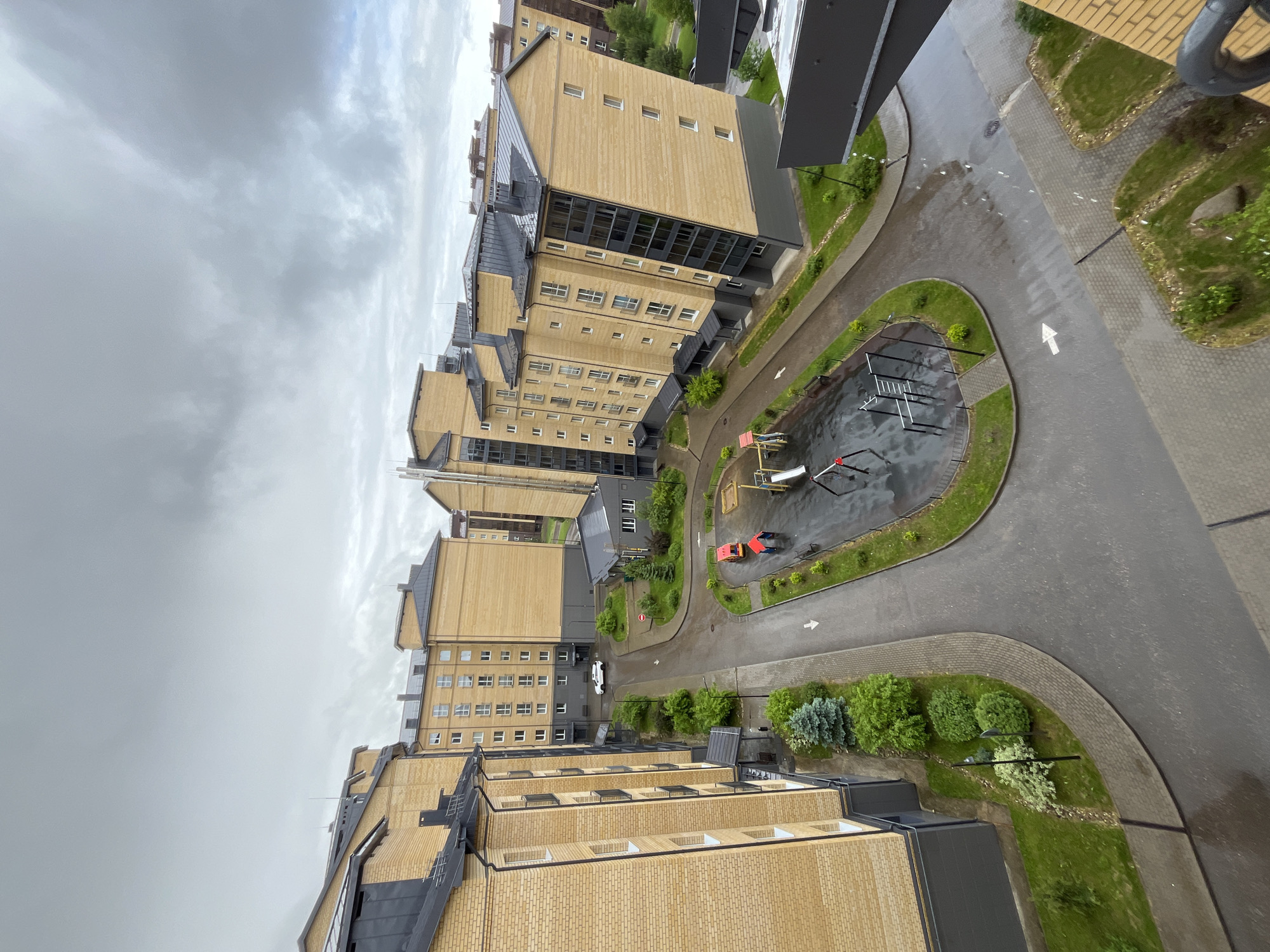 Here's a description of the image:
This is a high-angle shot looking down on a residential area in Мокшино, Russia. Several multi-story apartment buildings are visible, constructed of light brown brick with dark gray roofing and window accents. The buildings are arranged in a curved layout around a central courtyard area.
The courtyard contains a small playground with play equipment and red children's vehicle. Paved walkways and roadways wind through the courtyard, with neatly maintained green spaces featuring grass, shrubs, and small trees. White directional arrows are painted on the roadways.
The sky is overcast with gray clouds, suggesting a rainy or recently rainy day. The overall impression is of a modern, well-maintained residential complex. No people are visible in the image.