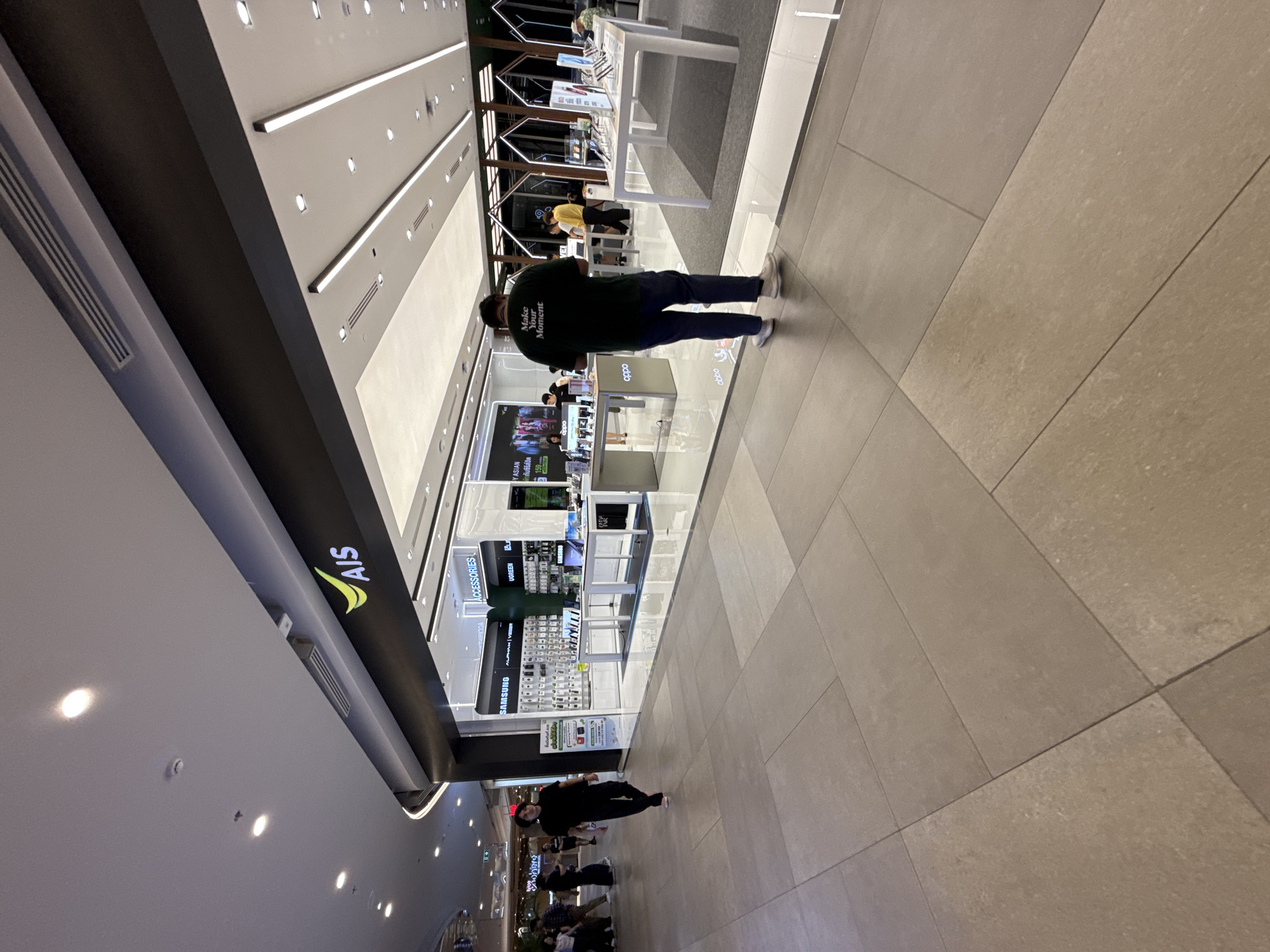 This media file captures an indoor scene within a brightly lit, modern retail environment, likely a shopping mall in Bangkok, Thailand. The primary subjects are several individuals shopping or working in an electronics store. One person in the foreground, seen from behind, wears a dark green t-shirt with text that reads "Make Your Moment." Another person, dressed in black, is also visible in the lower part of the frame.
The scene is set in a store that appears to sell mobile phones and accessories, indicated by prominent signage for "AIS," "SAMSUNG," "UGREEN," and "OPPO." Display shelves filled with products are visible, as are illuminated screens showing advertisements. The flooring is tiled in a neutral, light-colored stone. The lighting is bright and artificial, suggesting it is either evening or the interior is designed to be well-lit regardless of the time of day. The overall atmosphere seems busy and commercial, typical of a retail setting.
