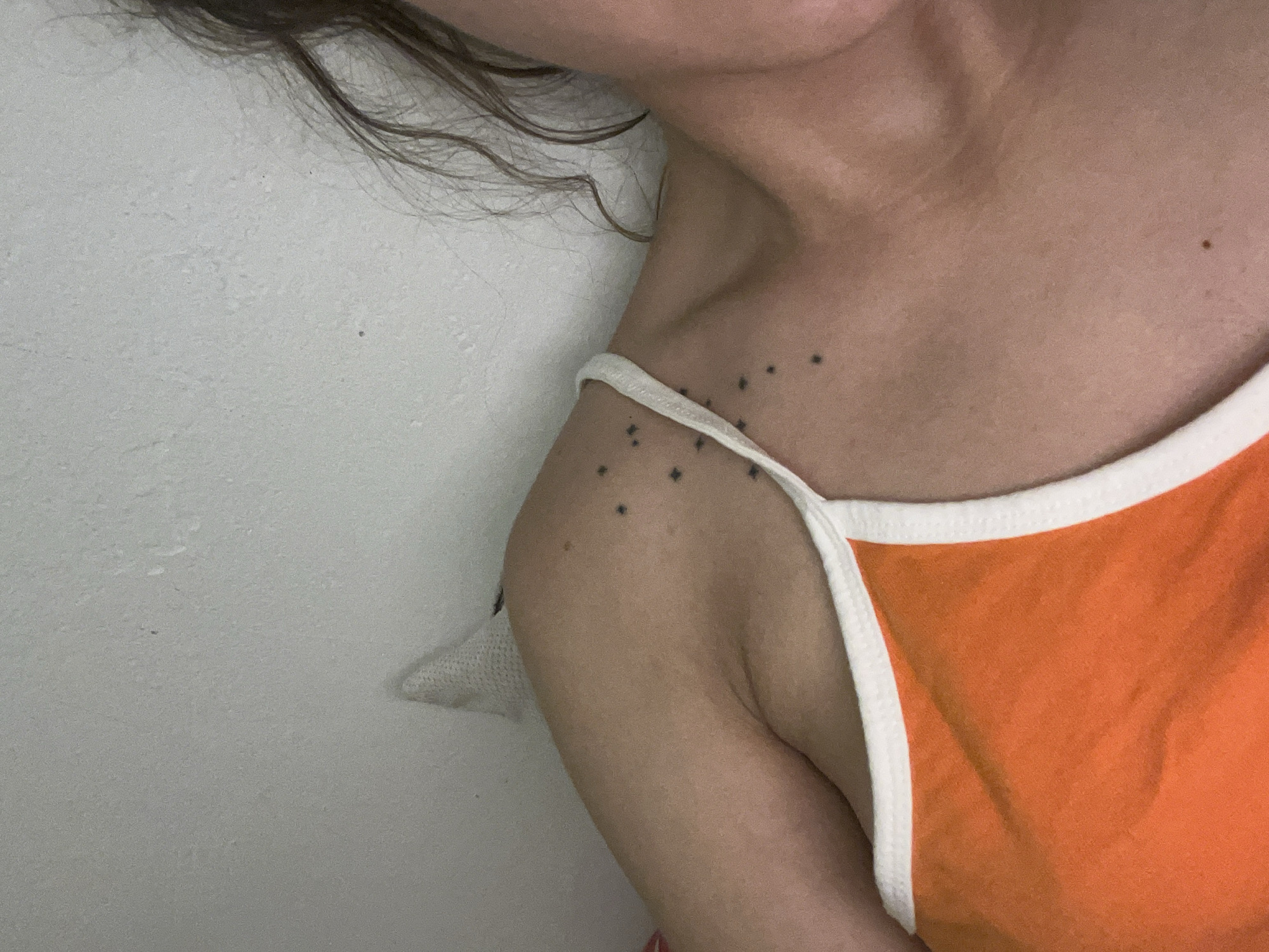A close-up photograph shows a partial view of a person's upper body. The person has fair skin and brown hair. The person is wearing an orange tank top with white trim. There are several small, black star-shaped tattoos on the person's shoulder. A mole is visible on the person's chest and shoulder. The background is a white wall. The image does not contain sufficient information to determine a specific location in Saarbrücken, Germany.