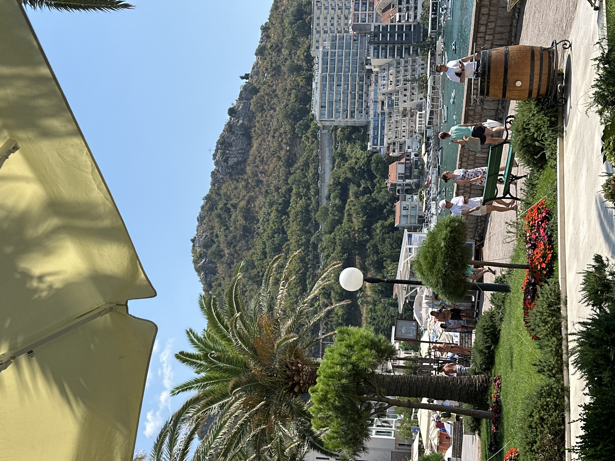 In Rafailovići, Montenegro, a sun-drenched scene unfolds along a waterfront promenade. A large, light yellow umbrella partially obscures the upper portion of the frame, casting shadows and creating a sense of shaded respite. Beneath it, a lush green lawn is dotted with vibrant red and pink flowers, bordered by dark green shrubs. A palm tree stands tall to the left, its fronds reaching towards a clear blue sky.
To the right of the lawn, a stone retaining wall separates the greenery from a paved walkway. Several people are present on the walkway. Four individuals, two women and two men, are visible. One woman, wearing a white cap and white t-shirt with khaki shorts, stands near a green park bench, holding a small white object. Beside her, another woman in a floral dress is seated on the bench. Further along the walkway, a third woman in a light green top and dark shorts carries a shopping bag. A man in a white t-shirt and dark shorts stands by a large wooden barrel, which appears to be decorative, holding a mobile phone. Behind the people, the calm, blue water of a bay is visible, dotted with numerous small boats.
In the background, a range of buildings forms a cityscape, extending up the hillside. These buildings include modern apartment blocks with glass balconies and older, more traditional structures with red-tiled roofs. The hillside behind the city is densely covered with trees and shrubs, culminating in a rocky peak topped with a cross. A road with parked cars is visible winding along the lower slope of the hill. A white lamppost with a spherical lamp stands on the walkway near the center of the scene. The overall atmosphere is one of a peaceful, sunny day at a coastal resort.