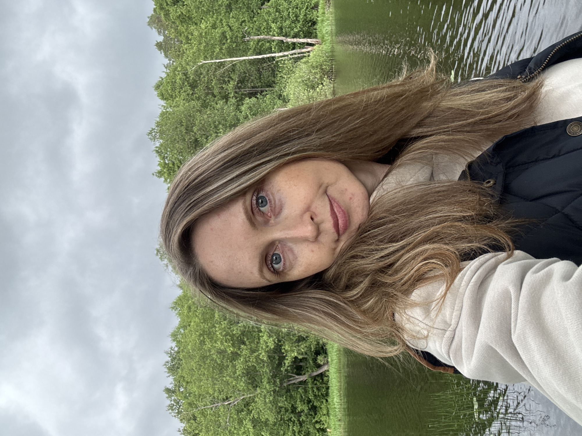 A female individual, appearing to be an adult, is captured in a close-up, slightly angled selfie, looking directly into the camera. She has long, wavy, light brown hair with blonde highlights, blue eyes, and visible freckles on her face. Her head is tilted slightly to her right, and she exhibits a faint, closed-mouth smile. She is wearing a light beige or oatmeal-colored hooded garment under a dark, possibly navy or black, quilted vest with metallic snap buttons. The background depicts a natural outdoor setting with a body of water, likely a lake or slow-moving river, reflecting the sky and surrounding trees. The far bank is densely covered with lush, vibrant green deciduous trees, suggesting late spring or summer, with a few lighter-colored bare tree trunks interspersed. The sky above is entirely overcast, featuring uniform grey clouds. The water is relatively calm with minor ripples and reflections. This scene is located in Chaikovsky, Russia.