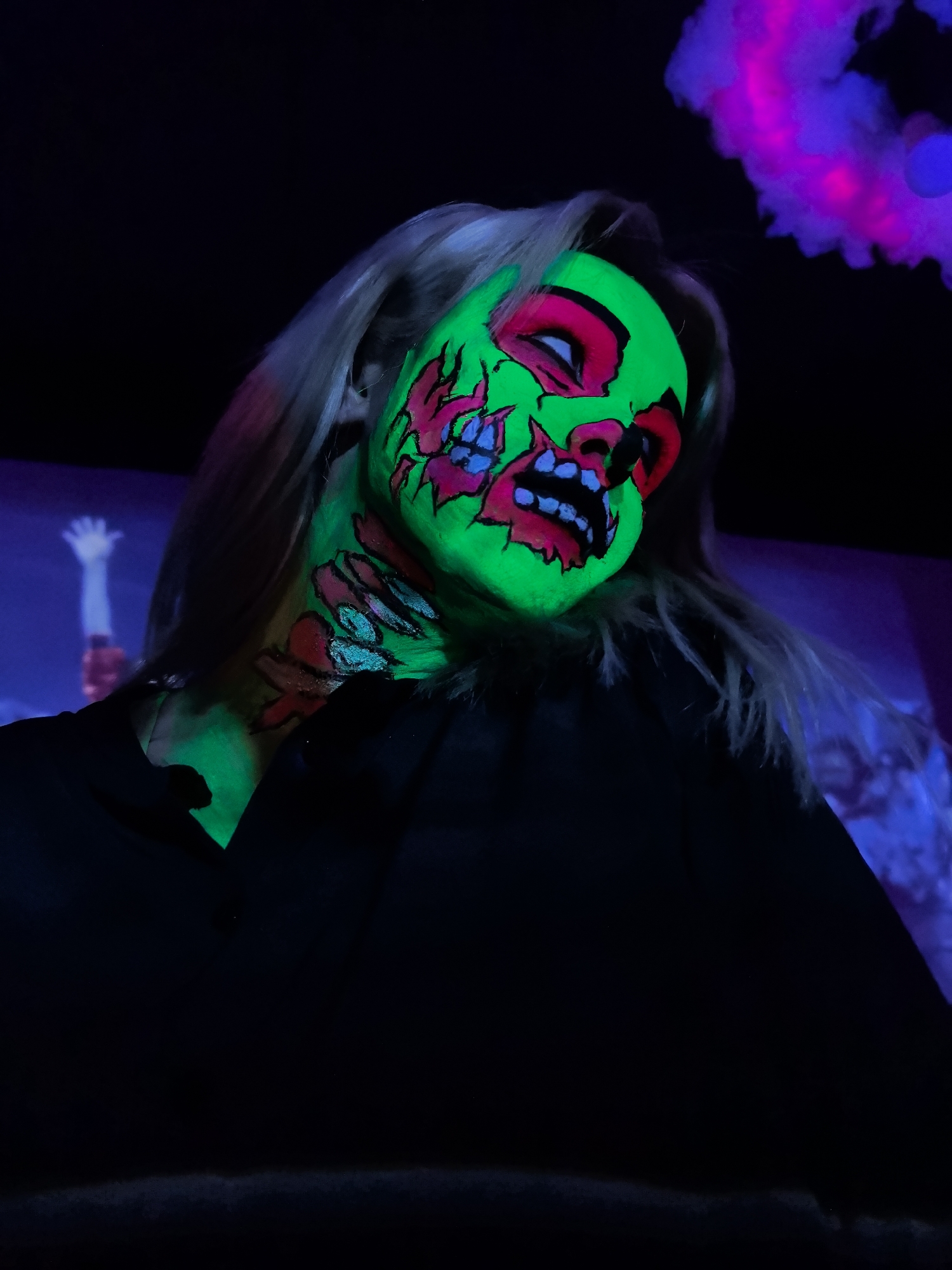 A close-up, low-angle, vertical shot depicts a person with elaborate face and neck makeup designed to resemble a zombie. The makeup is fluorescent and glows brightly under blacklight, with dominant colors of neon green, pink, and blue. The subject has fair skin and blonde hair, and is wearing a dark garment that obscures most of their body. The background is dark, with areas of blurry purple and blue light, suggesting an indoor setting with specialized lighting, possibly a club or event space. A faint, blurry image of a raised hand is visible in the mid-ground left, also illuminated by a purple light. The overall mood is eerie and theatrical. The location in the city of Noviy, Russia cannot be determined from the image.