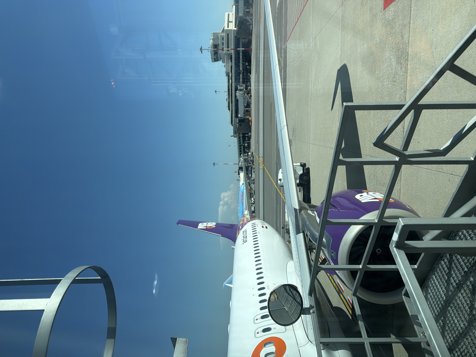 The image depicts an airport scene, likely taken from inside a terminal or an aircraft window. In the foreground, a portion of an airplane is visible, specifically its fuselage, wing, and engine. The aircraft has a white body with purple and orange accents, and the logo "AIR" is prominently displayed on the tailfin and engine.
Beyond the immediate aircraft, other planes are visible on the tarmac, including one with a blue and yellow livery. In the background, a large airport terminal building complex is situated, characterized by its modern architecture and control tower. The scene is set against a clear blue sky, suggesting fair weather. The airport environment includes a tarmac with clear markings and various airport vehicles.
The location is identified as Ferno, Italy, which is the municipality where Milan Malpensa Airport (MXP) is located. The image captures a typical operational moment at a major international airport. There are no discernible people in the image. The focus is on the aircraft, airport infrastructure, and the general ambiance of an airport on a sunny day.