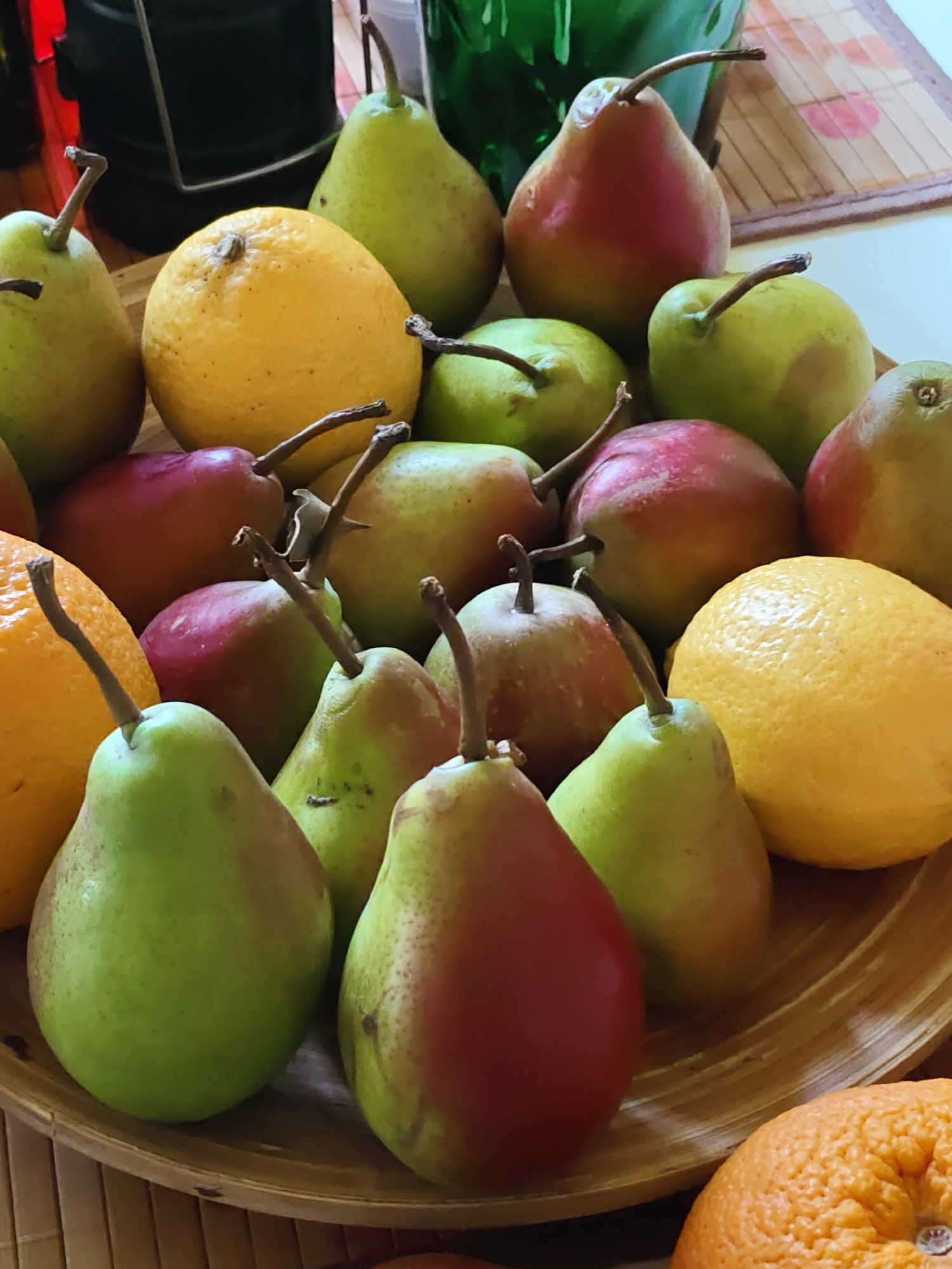 A wooden bowl is filled with a variety of fruits. The bowl rests on a woven placemat. The fruits include pears of various colors, including green, red, and yellow with red blush. There are also lemons and oranges. Some of the pears have stems attached. The image appears to be a still life, with no people or action depicted. The lighting is natural, casting soft shadows. The background is blurred, but suggests an indoor setting with some green objects visible.