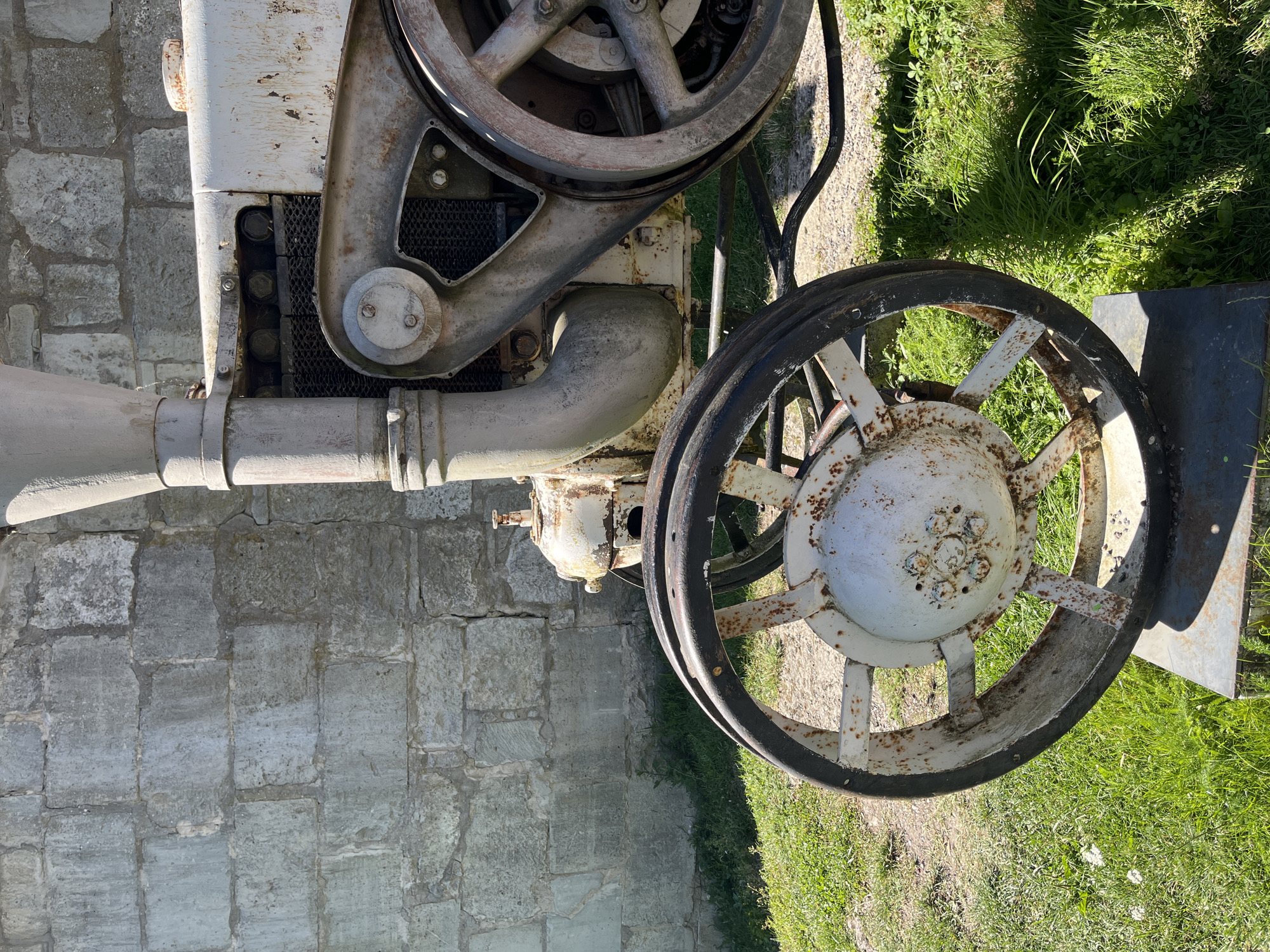 An antique, weathered white tractor with visible rust sits on display outdoors in front of a rough stone wall. The tractor's large, spoked wheels, one in the foreground with a black rim, are distinctive. The engine area shows a grate and various metal components. The environment is grassy with some sparse vegetation and gravel on the ground. The scene is illuminated by bright sunlight, casting shadows. The location is a historically significant area in Kuressaare, Estonia, specifically within the grounds of Kuressaare Castle, where old agricultural equipment is often displayed as part of the historical context of the region. There are no people visible in the image.