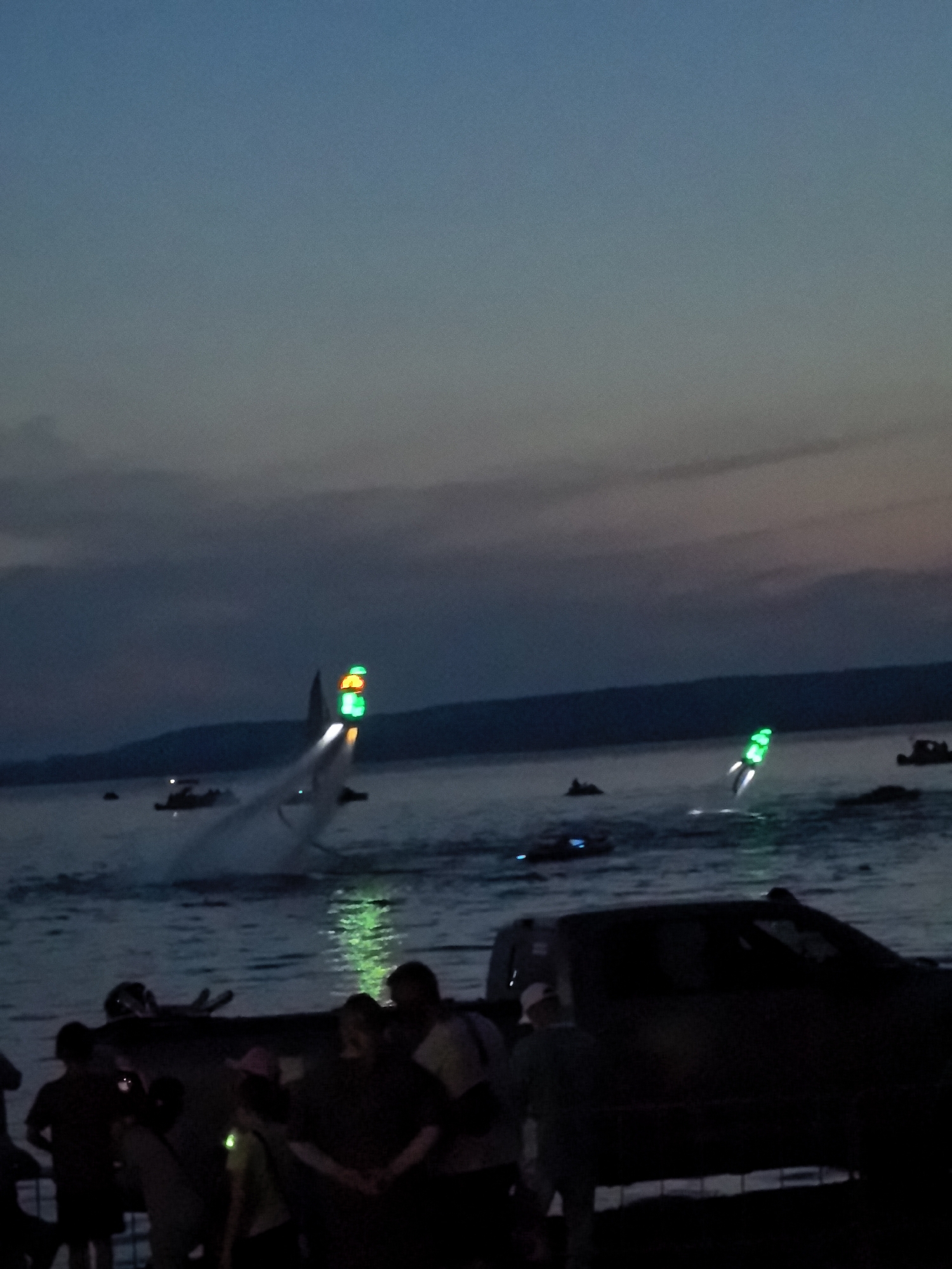 In the city of Osa, Russia, at dusk, a scene unfolds on the water's edge. A group of people are gathered on the shore, some silhouetted against the water, others standing near a dark-colored vehicle. In the distance, on the water, several boats are visible. One boat in particular is creating a significant wake and has a prominent, bright green light attached to its stern, as well as a smaller, orange light. Another boat further away also displays a bright green light. The sky above is a gradient of twilight blues and grays, with a faint hint of clouds. The overall impression is of a night-time event or gathering by the water.