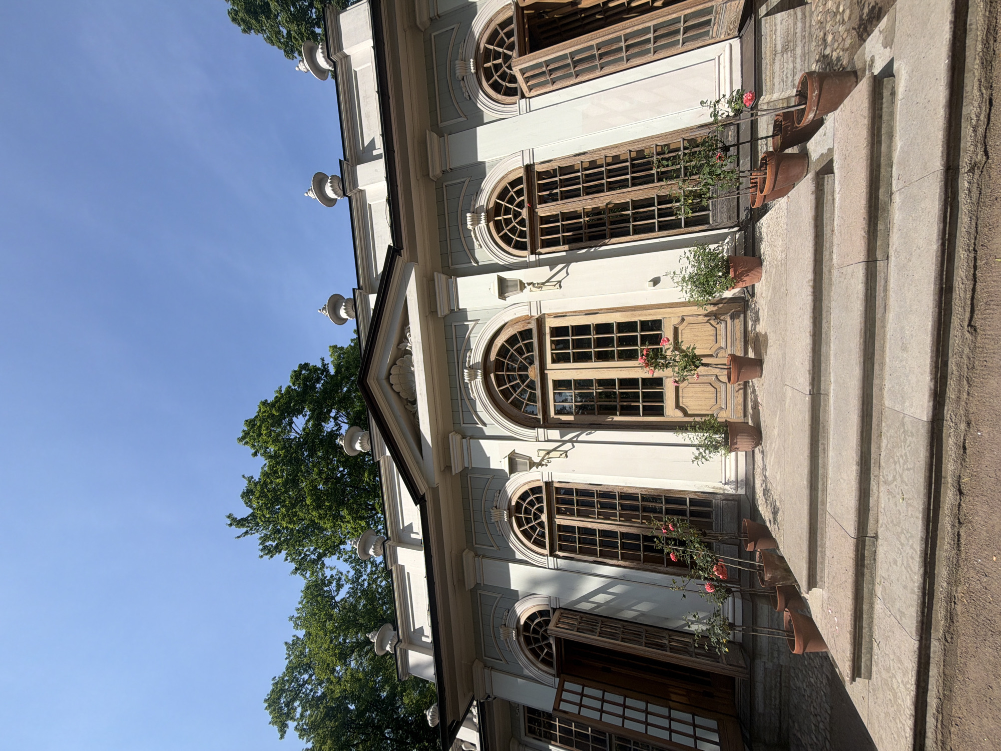 The image shows a white neoclassical pavilion with arched windows and doors, set against a backdrop of lush green trees and a clear blue sky. Several potted rose bushes are arranged on the stone steps leading up to the pavilion's entrance. The pavilion features ornate decorative elements on its facade and roofline, including finials and molding. Some of the wooden doors are open, revealing the interior structure. The scene suggests a serene outdoor setting, likely in a park or garden. Based on the architectural style and surrounding vegetation, this pavilion is likely located in the Summer Garden in Saint Petersburg, Russia, specifically the "Hermitage pavilion." The pavilion is a two-story structure with a colonnade on the ground floor and a loggia on the upper level, adorned with sculptures and bas-reliefs. It was built between 1719 and 1725.