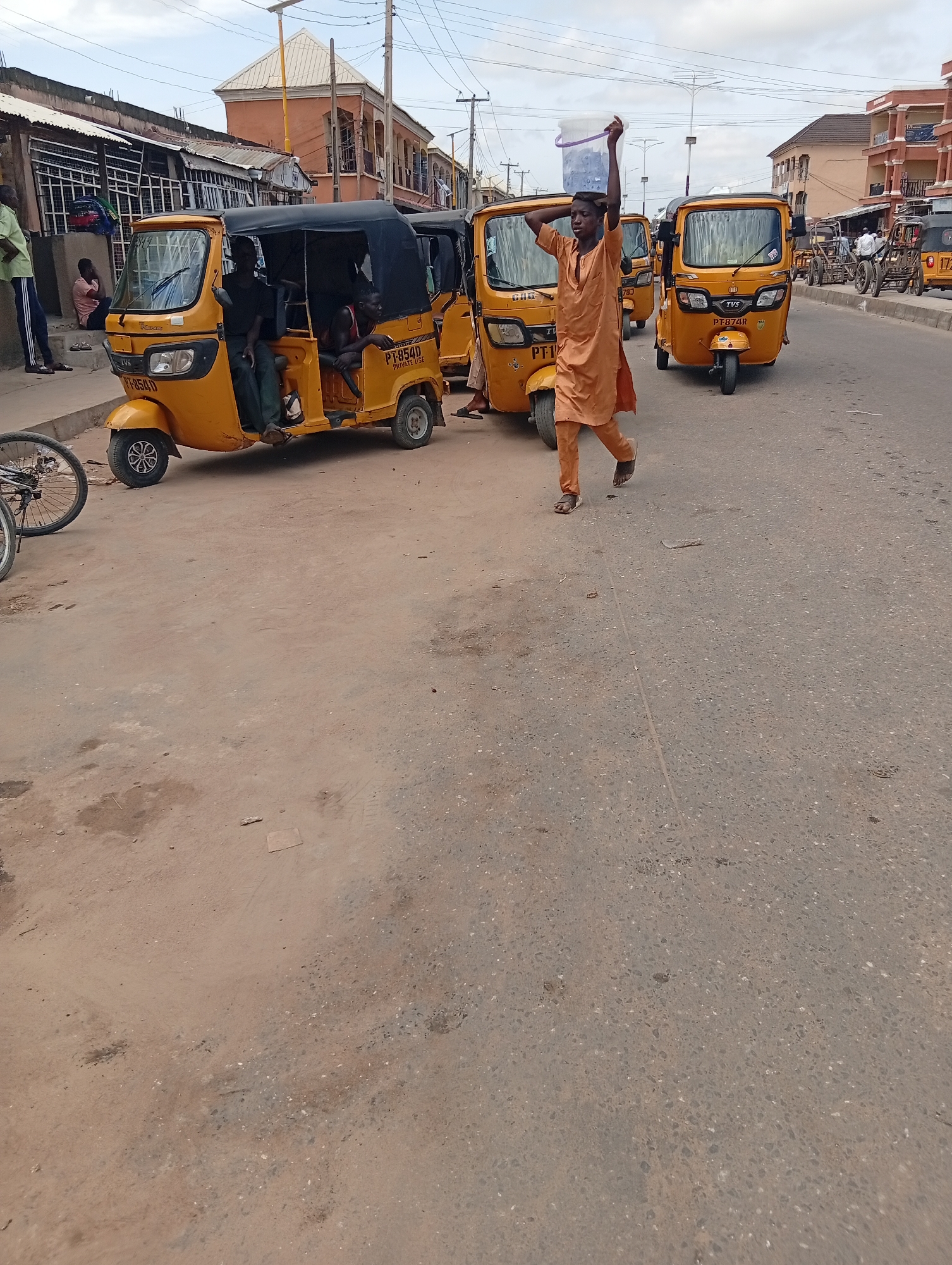 This image captures a bustling street scene in Jalingo, Nigeria, likely during the day given the bright lighting. The foreground is dominated by a dusty, paved road, with a parked yellow auto-rickshaw, commonly known as a "keke," on the left. Several other yellow kekes are visible further down the street, suggesting it's a common mode of transportation.
A young man in an orange outfit is walking in the middle of the street, carrying a large white bucket on his head with his right arm raised for balance. His posture and the determined look on his face suggest he is focused on his task. Other people are also present, with some seated in the kekes and others standing or walking in the background.
The background reveals a mix of buildings, some appearing to be shops or residences, with corrugated iron roofs and a few multi-story structures. Power lines are visible overhead, indicating the presence of electricity. The overall atmosphere seems to be one of everyday activity and commerce. The text visible on the kekes, such as "PT-8540" and "PT-874R," likely denotes registration numbers or route information. The scene depicts a typical day in a Nigerian urban environment, characterized by lively street life and the ubiquitous keke.