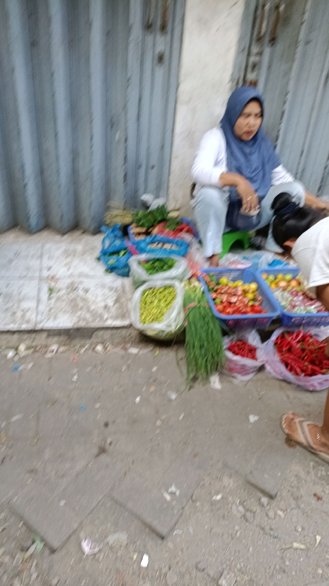 A woman wearing a blue hijab and white long-sleeved shirt sits on a green stool on the sidewalk in front of a closed metal roll-up door. She is looking to her right. In front of her, on the ground, are several plastic bags and blue plastic baskets filled with produce, including green beans, red chilies, tomatoes, and onions. Another person, wearing a white shirt, is partially visible on the right, bent over and looking at the produce. The ground is concrete, with some debris. The scene appears to be a street market stall in Surabaya, Indonesia.