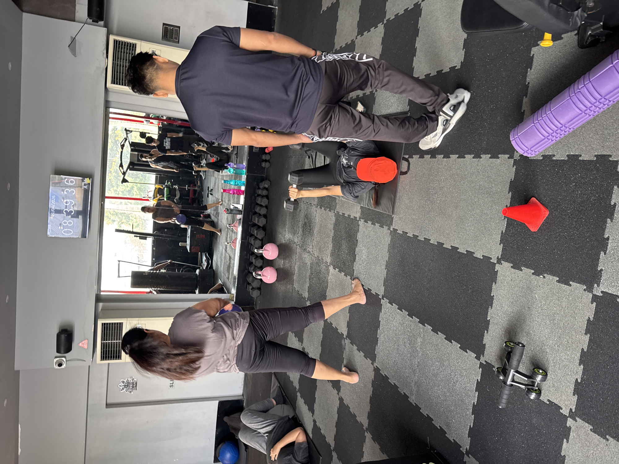 The image shows the interior of a fitness center in Kolkata, India. The floor is covered with gray and black interlocking rubber mats. There are multiple individuals within the frame. In the foreground, a person in black athletic pants and no shoes is performing a workout, seemingly a side lateral raise with what appears to be a kettlebell. Above them, a person in a dark-colored t-shirt and athletic pants with white stripes is standing and appears to be monitoring or assisting another person lying on a weight bench, who is doing a dumbbell press while wearing an orange cap. There are pink kettlebells on the floor near the mirror. The gym equipment is visible in the reflection of a mirror along one wall. A digital clock on the wall displays what appears to be the time and date as 08:39:36 and 21/04/2026. A purple foam roller and a red cone are situated near the weight bench, while an ab roller is on the floor in the bottom right corner.