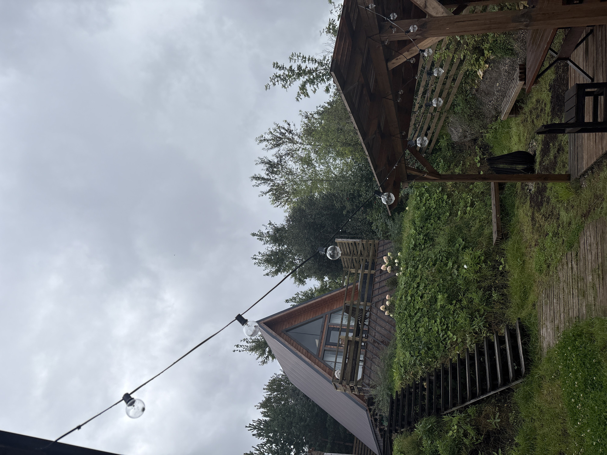 The image depicts a wooden A-frame cabin situated on a hillside surrounded by trees and lush greenery. A wooden staircase leads up the hill towards the cabin, with a wooden pathway at its base. String lights with exposed bulbs are strung across the scene, illuminating a wooden gazebo or covered seating area in the foreground, which includes a table and chairs. The sky is overcast. The location is the city of Chaikovsky, Russia.