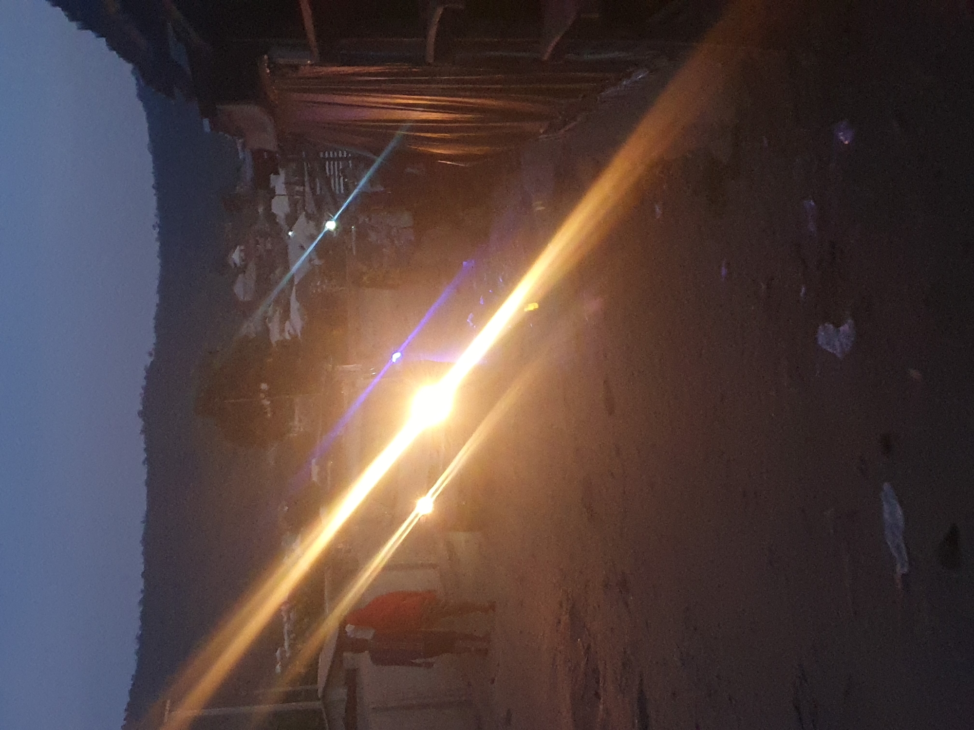 At dusk in Lokoja, Nigeria, a street scene is illuminated by a bright light, which reflects off the dark road surface. A person wearing a red shirt stands near a light-colored structure. To the left, a dark mountain range is visible under a blue sky. Overhead, there is a wooden structure. The road shows signs of debris.