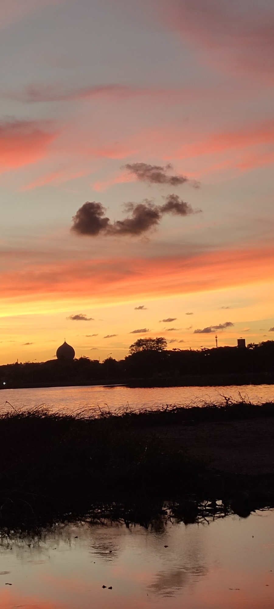 This is a vertical, eye-level, medium shot of a sunset over a body of water, likely a river or lake, in Lhokseumawe, Indonesia. The sky is painted with vibrant hues of orange, pink, and yellow, with streaks of darker clouds scattered throughout. The sun has set, casting a warm glow that reflects beautifully on the still water. In the background, silhouetted against the colorful sky, is a distinct domed building, possibly a mosque, and other buildings and trees lining the horizon. The foreground is dominated by dark, silhouetted vegetation and the water's surface, which also mirrors the sky's colors and the surrounding elements. The overall mood is serene and peaceful, capturing the tranquil beauty of a tropical sunset. The time of day is clearly sunset, indicated by the colors in the sky and the silhouette effect. There is no visible text in the image.