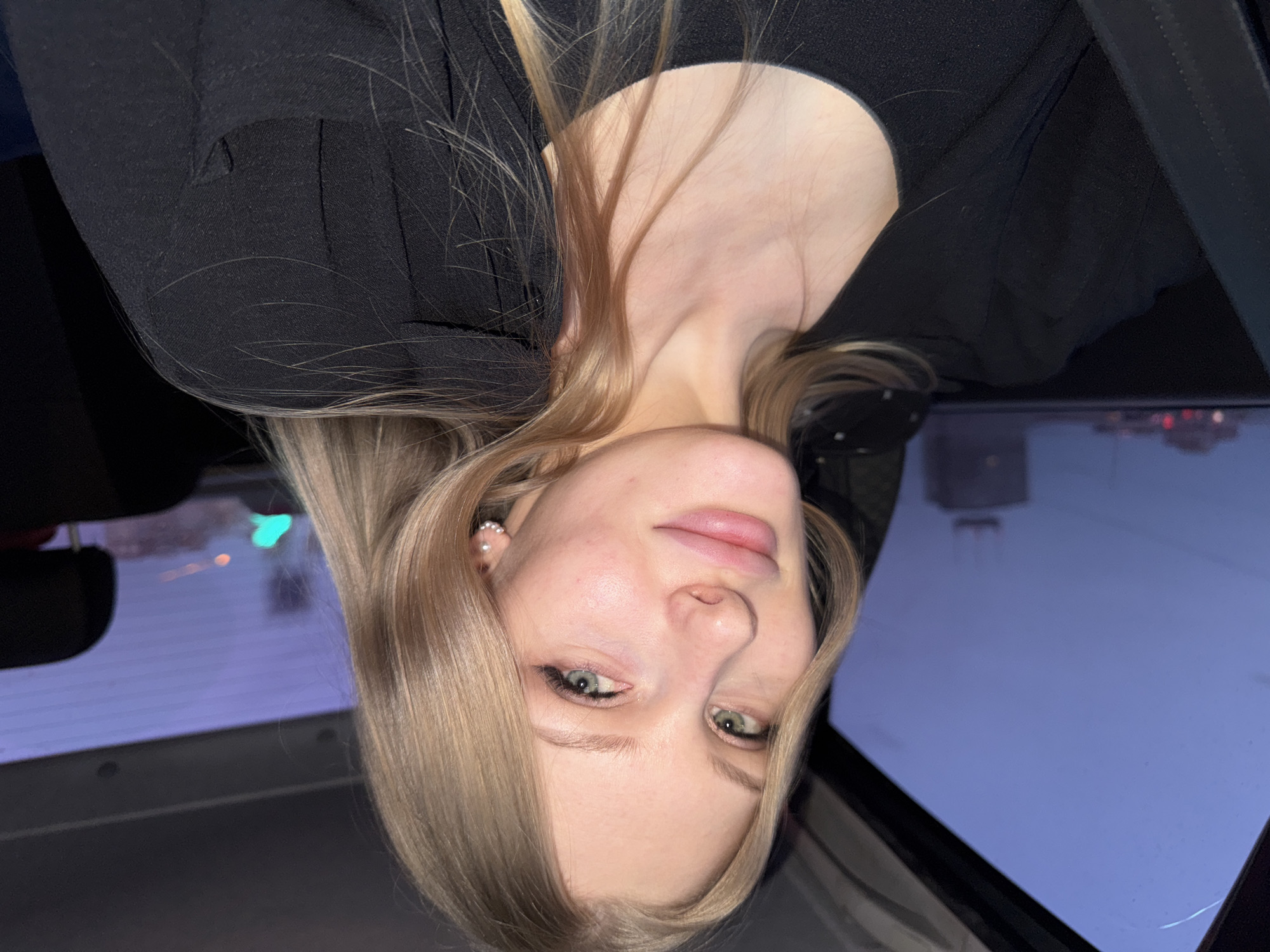 A blonde woman with green eyes is in the front seat of a car. She is wearing a black jacket or top. Outside the car window, there is a glimpse of the cityscape of Perm, Russia with various buildings and streetlights visible. The image is taken from an unusual angle, as if the camera were positioned upside down.