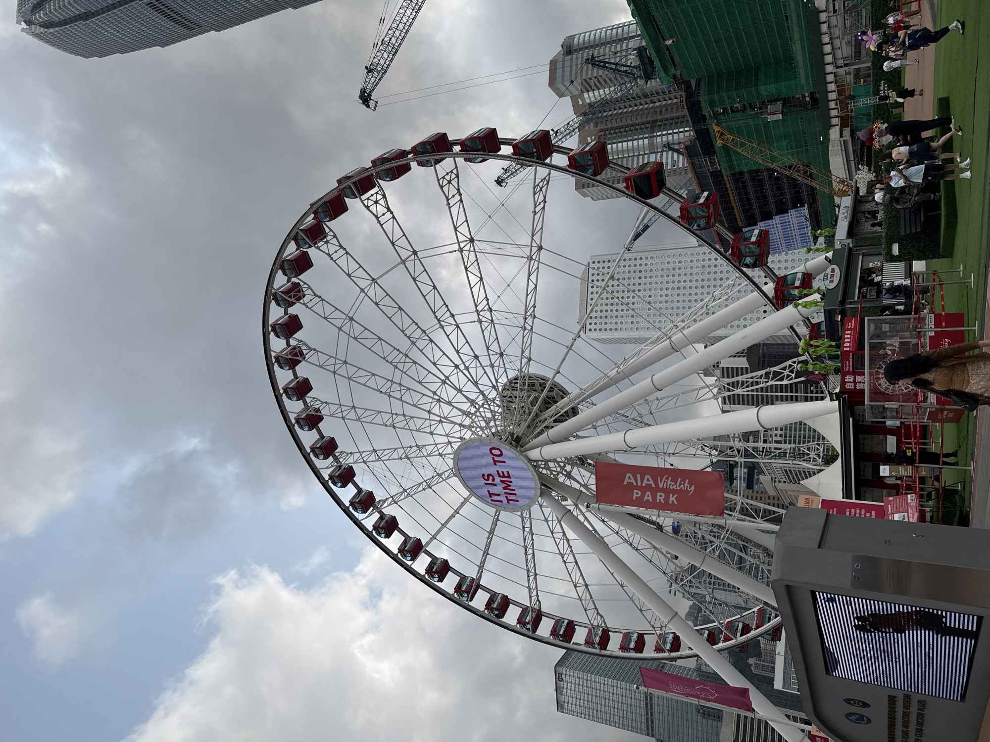 Here's a detailed description of the image:
The image captures a Ferris wheel in Hong Kong, China. The wheel, painted white with red gondolas, is the central focus. A large circular sign at the hub reads "IT IS TIME TO". A banner below reads "AIA Vitality PARK".
In the background, modern skyscrapers rise, reflecting the urban setting of Hong Kong. Construction cranes are visible amongst the buildings, indicating ongoing development. The sky is overcast with gray clouds.
On the right side of the image, there's a small green area, and some people are standing and walking. Signage and display boards are visible in the foreground, partially obscuring the view of the ground-level activity.