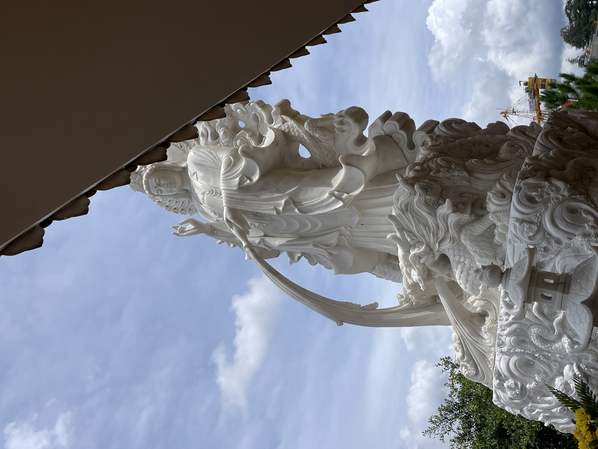 The image shows a large white marble statue of a female deity, possibly Guan Yin, set against a cloudy blue sky. The statue is intricately carved, with flowing robes and decorative elements that appear to be dragons. The deity is depicted in a standing pose, with one hand raised and the other holding a stream of water. The statue is mounted on a complex base that also features dragon motifs and a small pagoda-like structure. In the background, to the right, a yellow construction crane is visible, suggesting ongoing development in the area. To the left, lush green trees are present. The statue is likely located at the Linh Phuoc Pagoda in Da Lat, Vietnam, a well-known landmark famous for its intricate Buddhist statuary and mosaic artwork.