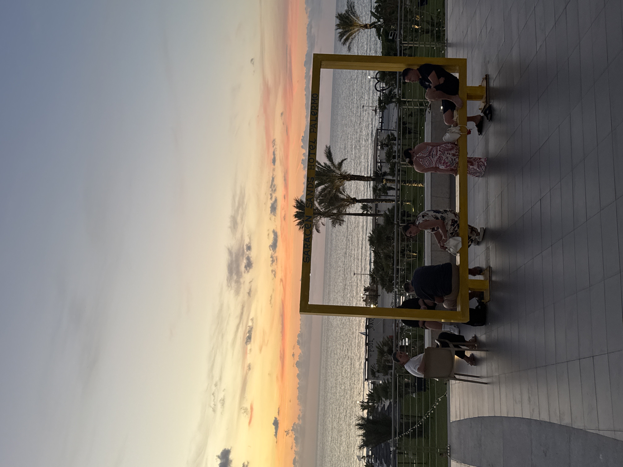 At sunset, seven individuals are gathered around a large, bright yellow, rectangular frame structure. The structure is positioned on a paved outdoor area overlooking the sea. The sky displays a gradient of colors, from soft oranges and pinks near the horizon to a pale blue overhead, with scattered clouds. The sea is calm, with a few palm trees visible along the coastline in the middle ground. The yellow frame has text printed on its top beam, which appears to read "SARACEN SANDS HOTEL PALERMO".
The individuals are seated or leaning against the frame. On the far left, a man in a white shirt and dark pants is sitting on a chair. Next to him, a woman in a patterned dress is seated. To her right, a woman in a floral dress is seated. In the center, a man in a dark shirt and shorts is seated. To his right, a woman in a patterned dress is seated. On the far right, a man in a black t-shirt and shorts is seated, leaning against the frame. All individuals appear to be casually dressed and are facing various directions. In front of the frame, a black backpack is placed on the ground. The paved area consists of dark gray square tiles. A railing runs along the edge of the paved area.