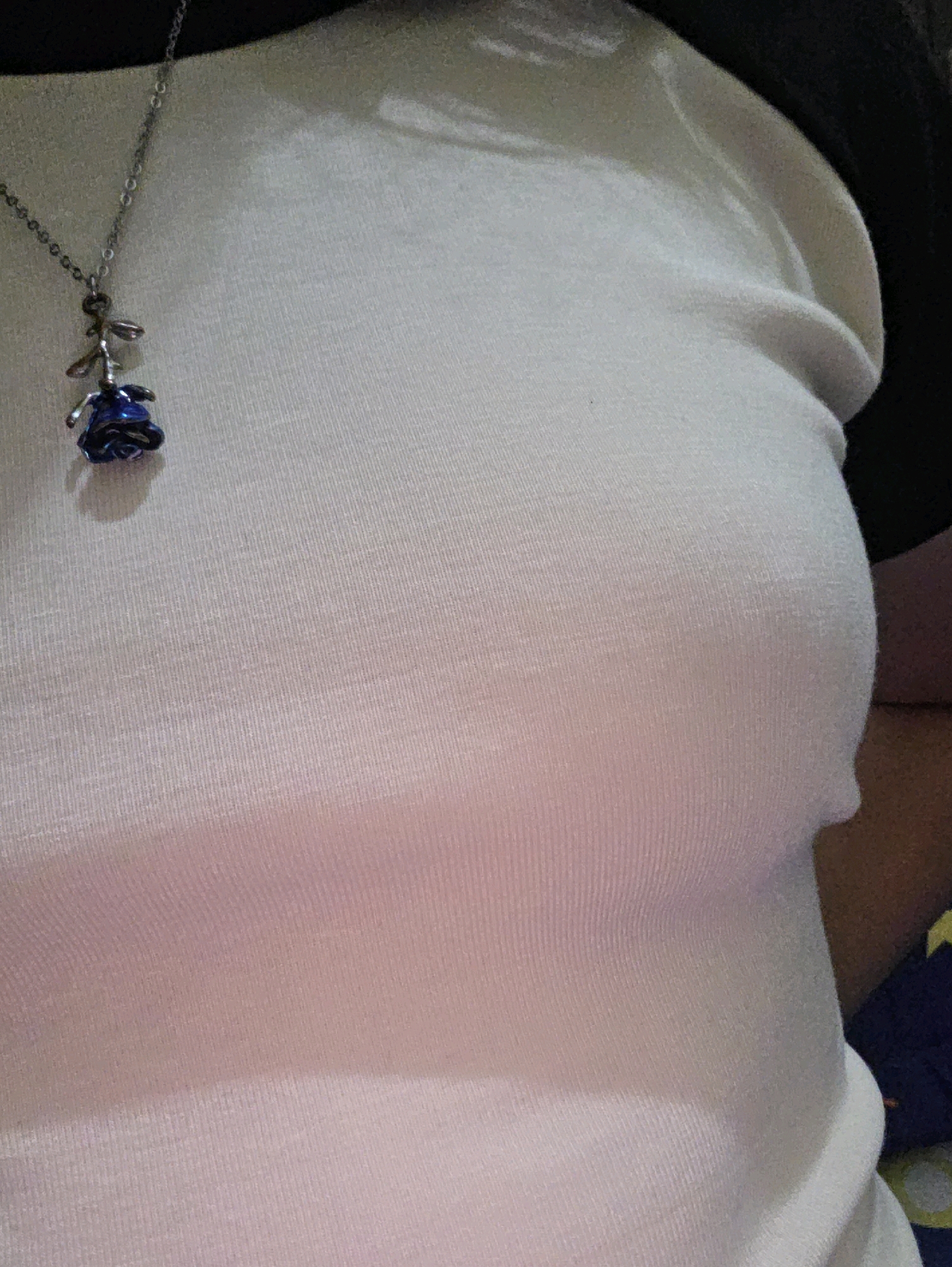 The image shows a close-up of a person wearing a white t-shirt and a necklace with a blue rose pendant. The lighting is dim, creating shadows on the fabric. The background is dark, with a hint of a patterned fabric in the lower right corner. There are no discernible city landmarks or features in the image, and therefore, it is not possible to determine its location within Phan Thiết, Vietnam.