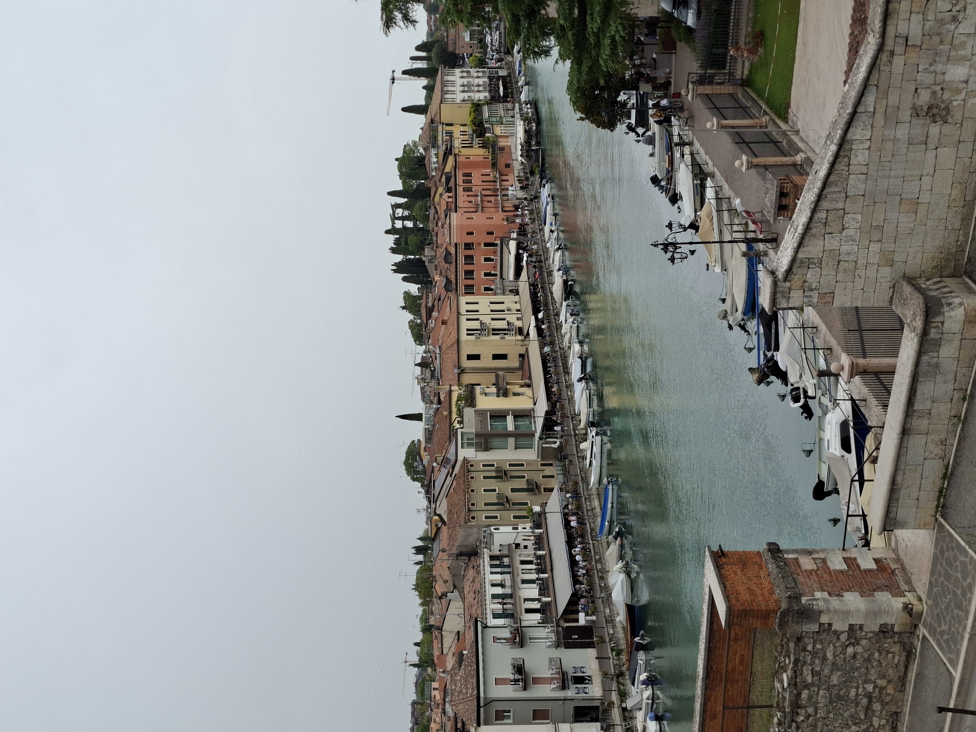 The image depicts a view of the canal in Peschiera del Garda, Italy, with buildings lining the waterfront and numerous boats docked along the side. The canal is calm and reflects the overcast sky, which appears to be a pale gray.
On the left side of the canal, a row of multi-story buildings with terracotta roofs are visible. These buildings house shops and restaurants at street level, indicated by awnings and tables set up outdoors where people are seated. The architecture is varied, with some buildings appearing older with traditional facades and others more modern. Balconies and windows are visible on the upper floors.
Along the canal's edge, a walkway is lined with benches and lampposts. Several small boats, mostly motorboats, are moored along the walkway. Some are covered with tarps, while others are uncovered. The water is a light blue-green color.
On the right side of the canal, more buildings and trees are visible. A pathway runs alongside the canal, with people walking and a car parked on the far right. The scene suggests a lively waterfront area, likely a tourist destination.
In the foreground, a stone wall and a brick structure, possibly part of a historical fortification or a scenic overlook, frame the view of the canal. The ground in the foreground is paved with stone and gravel pathways. The overall impression is one of a charming European town with a prominent waterway.