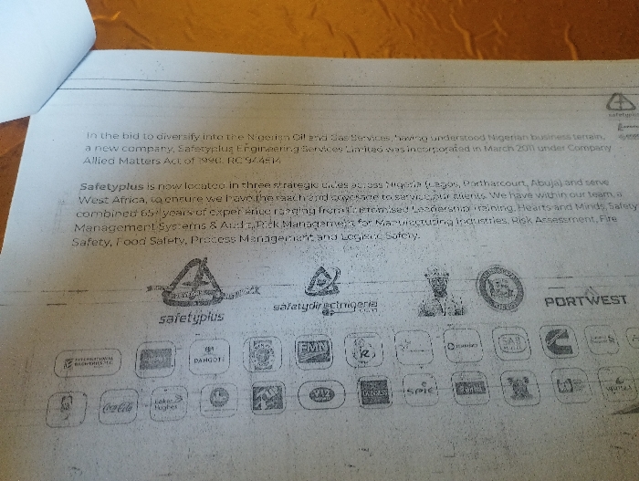 The image is a close-up photograph of a white sheet of paper with printed black text. The paper is slightly wrinkled and the print is faded. The text is an advertisement for a company called Safetyplus Engineering Services Limited, which is located in Ashaka, Nigeria (latitude: 10.917, longitude: 11.475). The advertisement describes Safetyplus as a new company that was incorporated in March 2011 under the Nigerian Company Allied Matters Act of 1990, RC 944514. Safetyplus offers services across Nigeria (Lagos, Port Harcourt, Abuja) and West Africa, including safety training, leadership training, risk assessment, fire safety, food safety, process management and hygiene safety. The text contains a variety of logos of other companies that are clients of Safetyplus. There are no people or objects in the image besides the paper with the advertisement. The environment is a close-up of a printed page.
