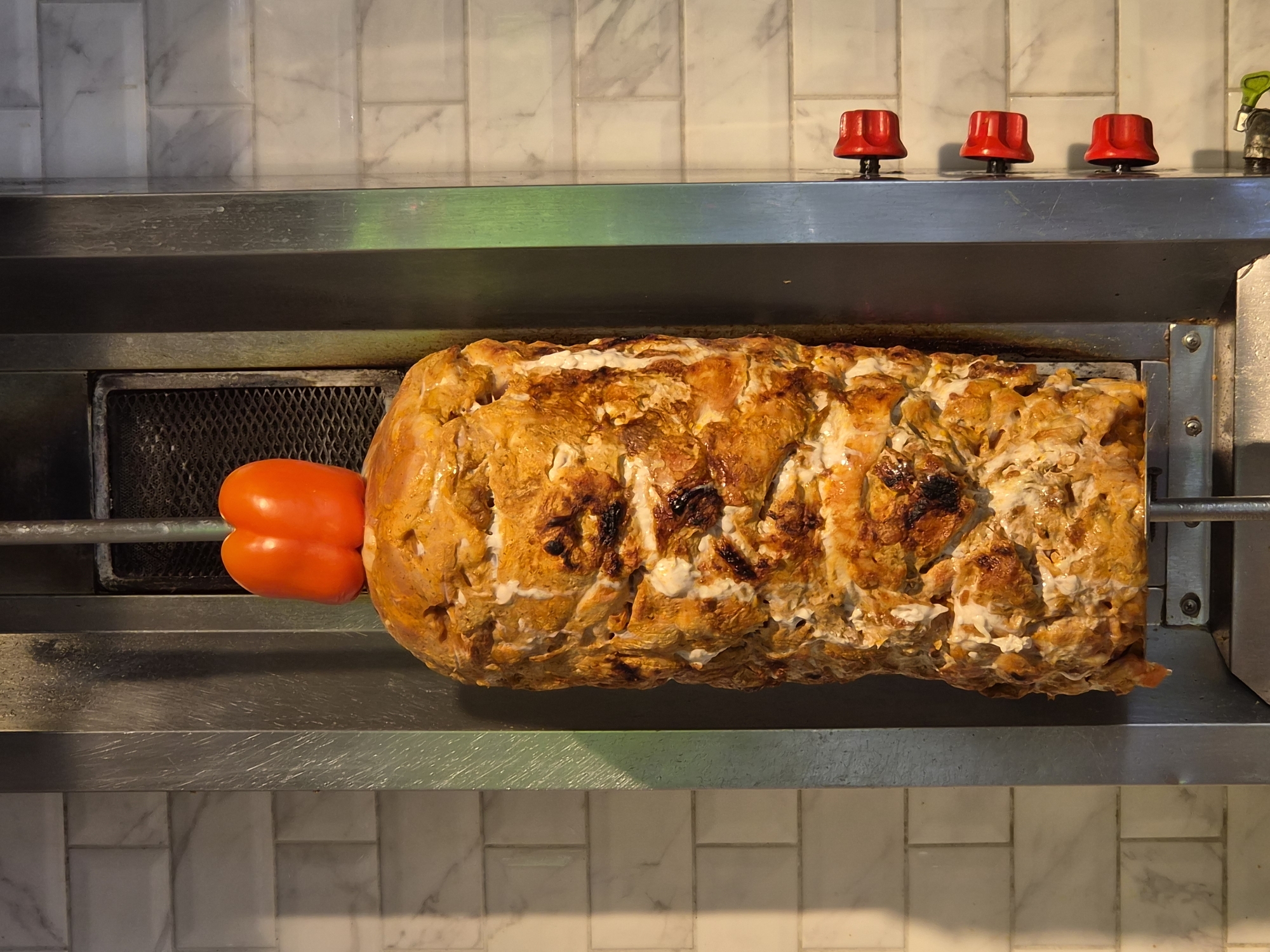 A front-facing medium shot displays a metal apparatus holding what appears to be a large, cylindrical mass of cooked meat and a red bell pepper. The metal apparatus is mounted on a tiled wall, giving the impression of a kitchen environment. The meat is a mixture of white and orange shades, with some charred sections. The bell pepper is positioned at the edge of the meat mass. At the top of the metal apparatus are three red knobs, seemingly controls for the apparatus. The overall impression is that of food preparation, possibly shawarma or a similar dish, in a restaurant or food service setting. There is no information available in the photo to locate the scene specifically in the city of Sochi, Russia.